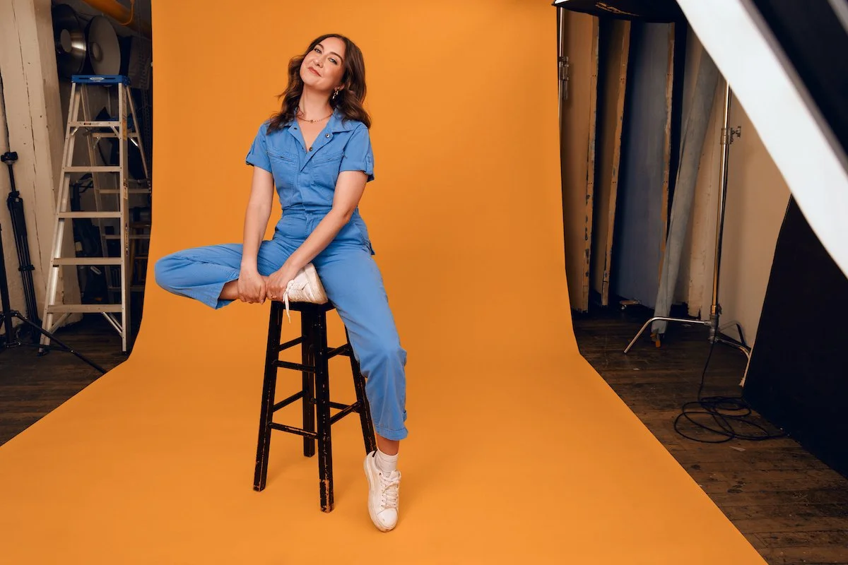 A woman wearing a blue jumpsuit and white sneakers sitting on a black stool against an orange backdrop in a photography studio.