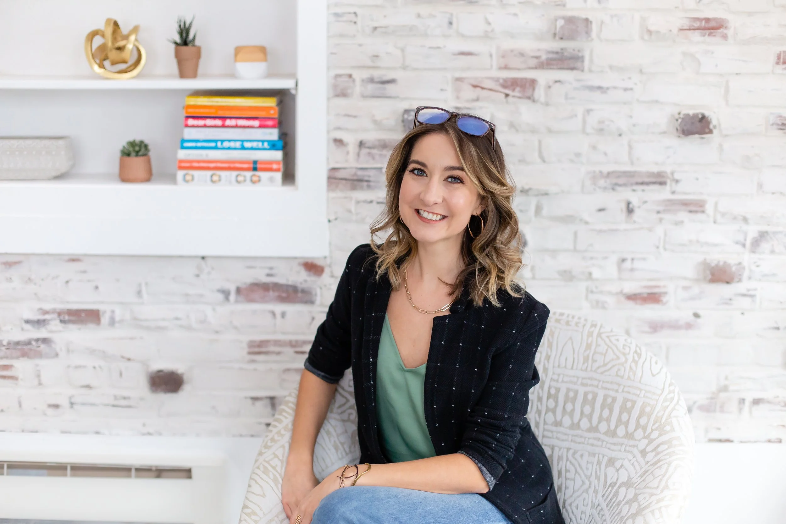 A smiling woman with wavy blonde hair and glasses on her head, sitting on a couch in a modern room with white brick walls, a white bookshelf with colorful books, and decorative items.