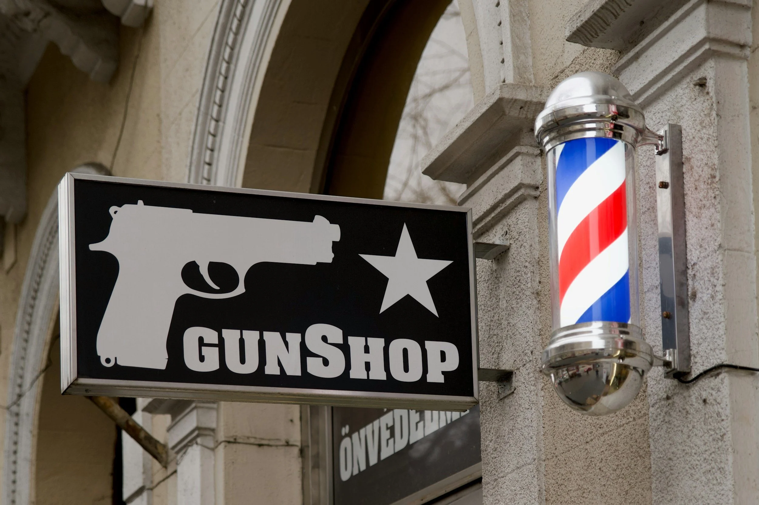 A store sign for a gun shop featuring a white pistol silhouette, a star, and the text 'GUNSHOP' in white letters on a black background, mounted on a beige brick wall with a traditional barber pole light fixture nearby.
