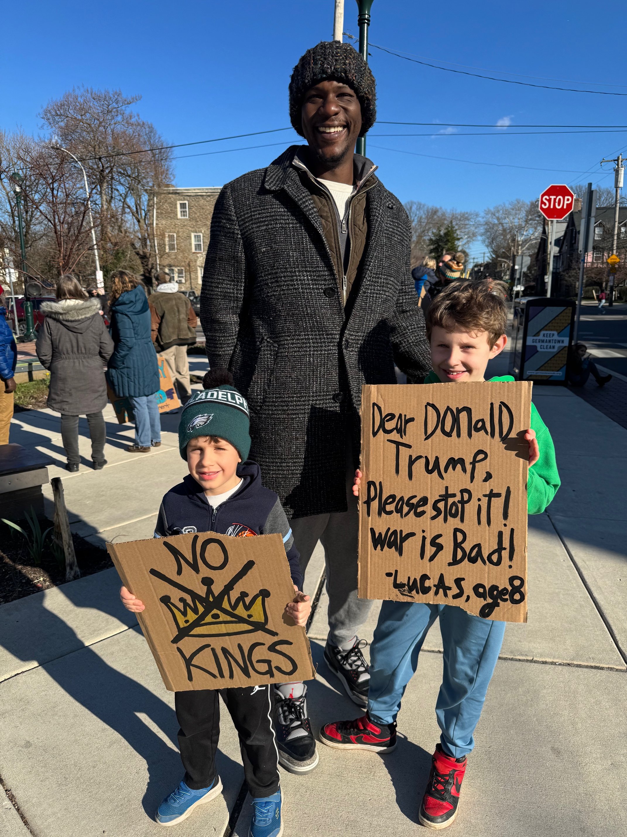 Two children and an adult man holding protest signs on a sidewalk during a demonstration. The boy on the left is holding a sign that says 'NO KINGS' with a crown drawing, and the boy on the right is holding a sign that says 'Dear Donald Trump, Please stop it! war is bad! -Lucas, age 8'. The man is smiling, and there are other protesters in the background with signs, trees, buildings, and a clear blue sky.