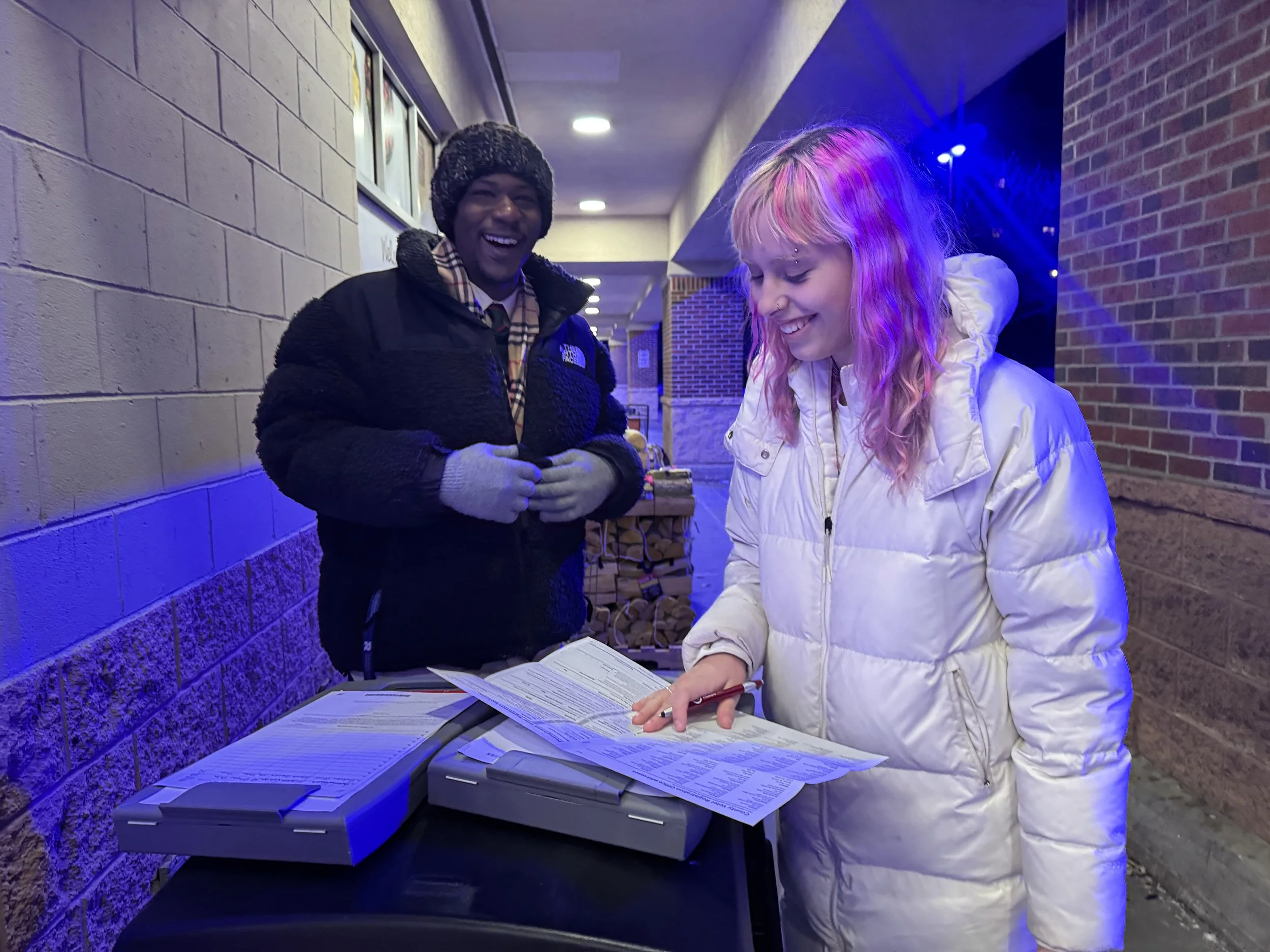 A young woman in a white puffer jacket looking at a menu or flyer at a counter, smiling, while a smiling man in a black jacket and beanie stands nearby in an outdoor setting under blue lighting.