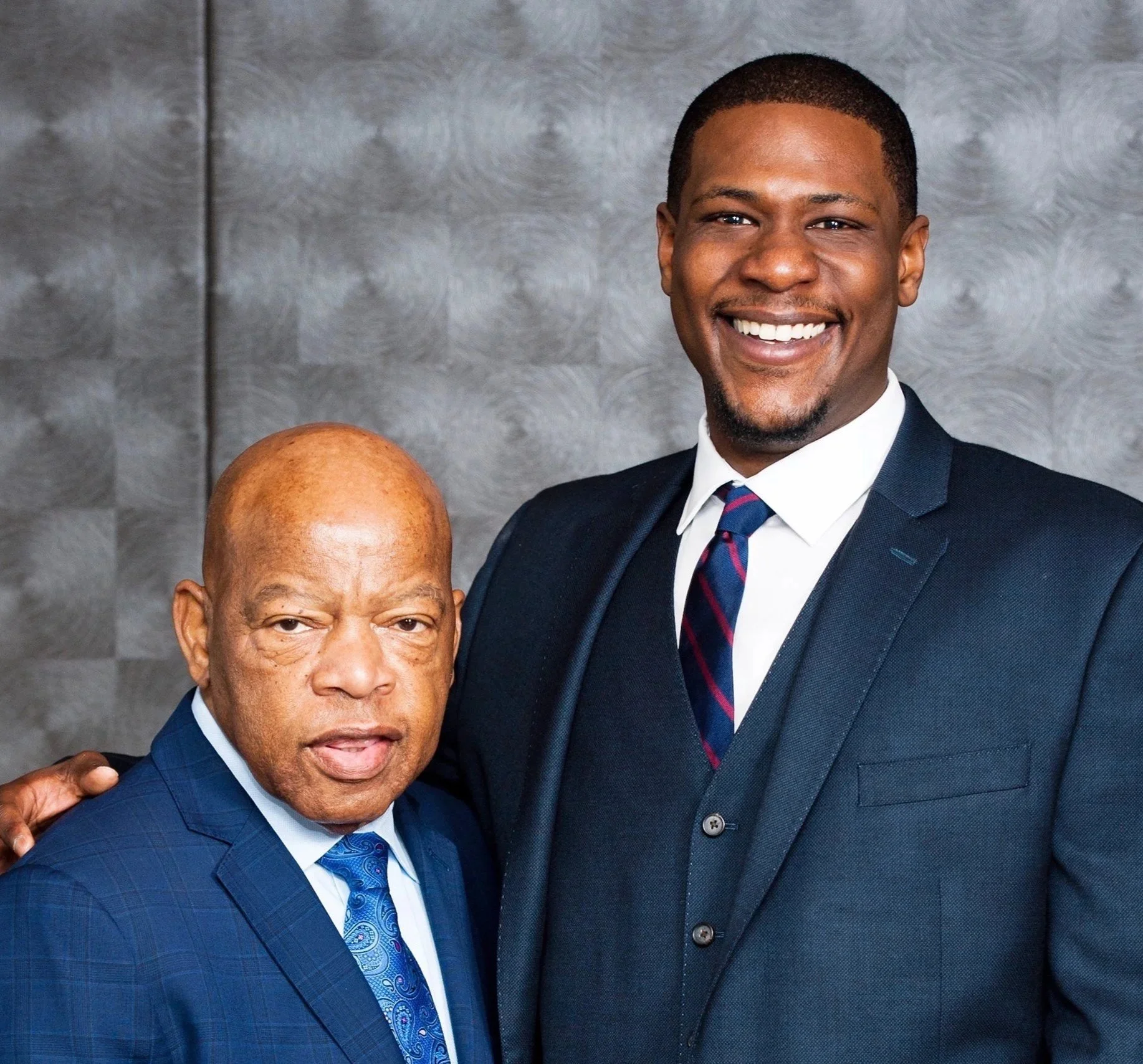 Two men dressed in suits smiling at the camera, standing against a gray textured background. The man on the left is older with a bald head, and the man on the right is younger with short hair.