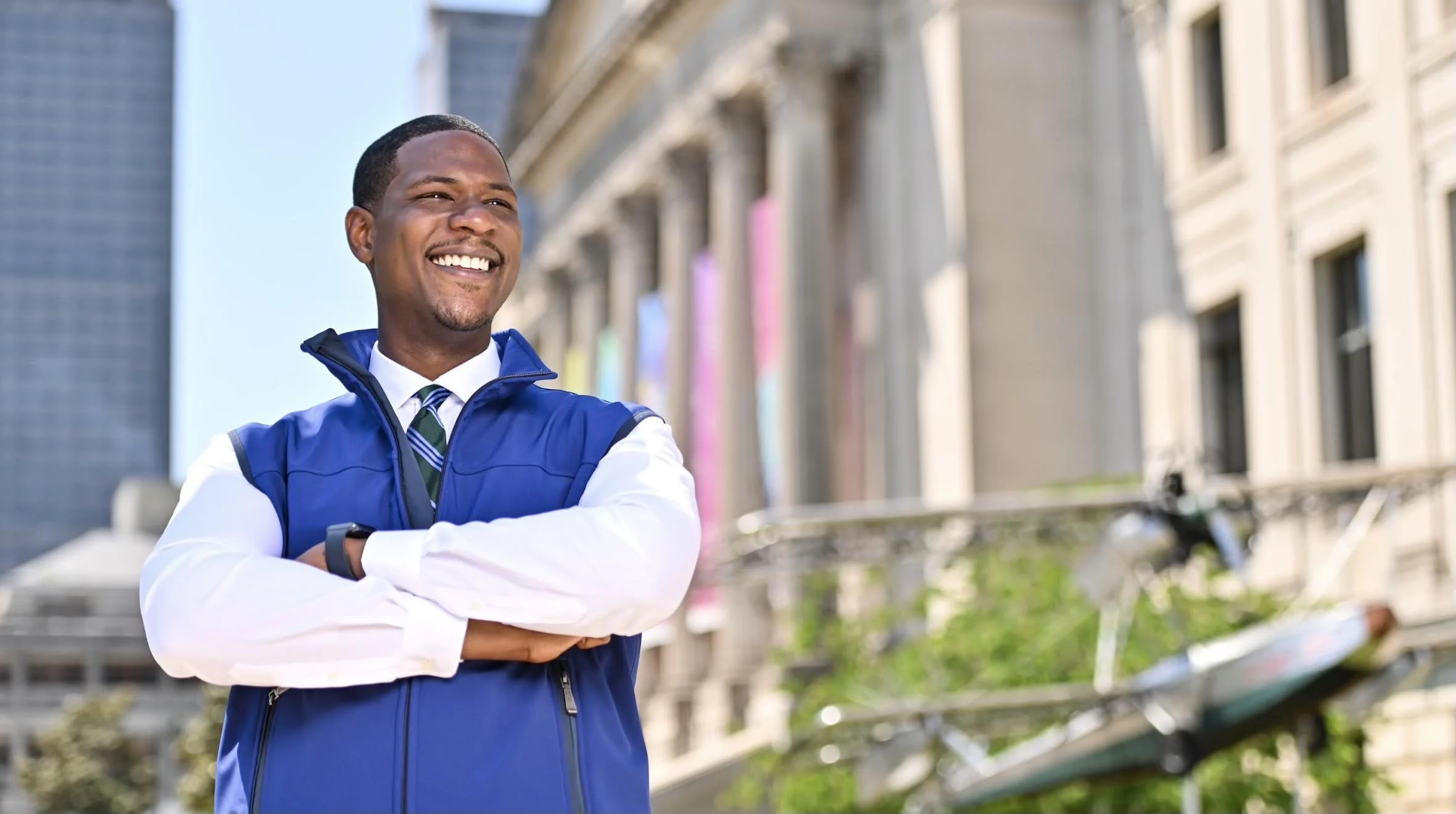 Smiling man wearing a blue vest and white shirt standing outdoors in front of buildings with arms crossed.