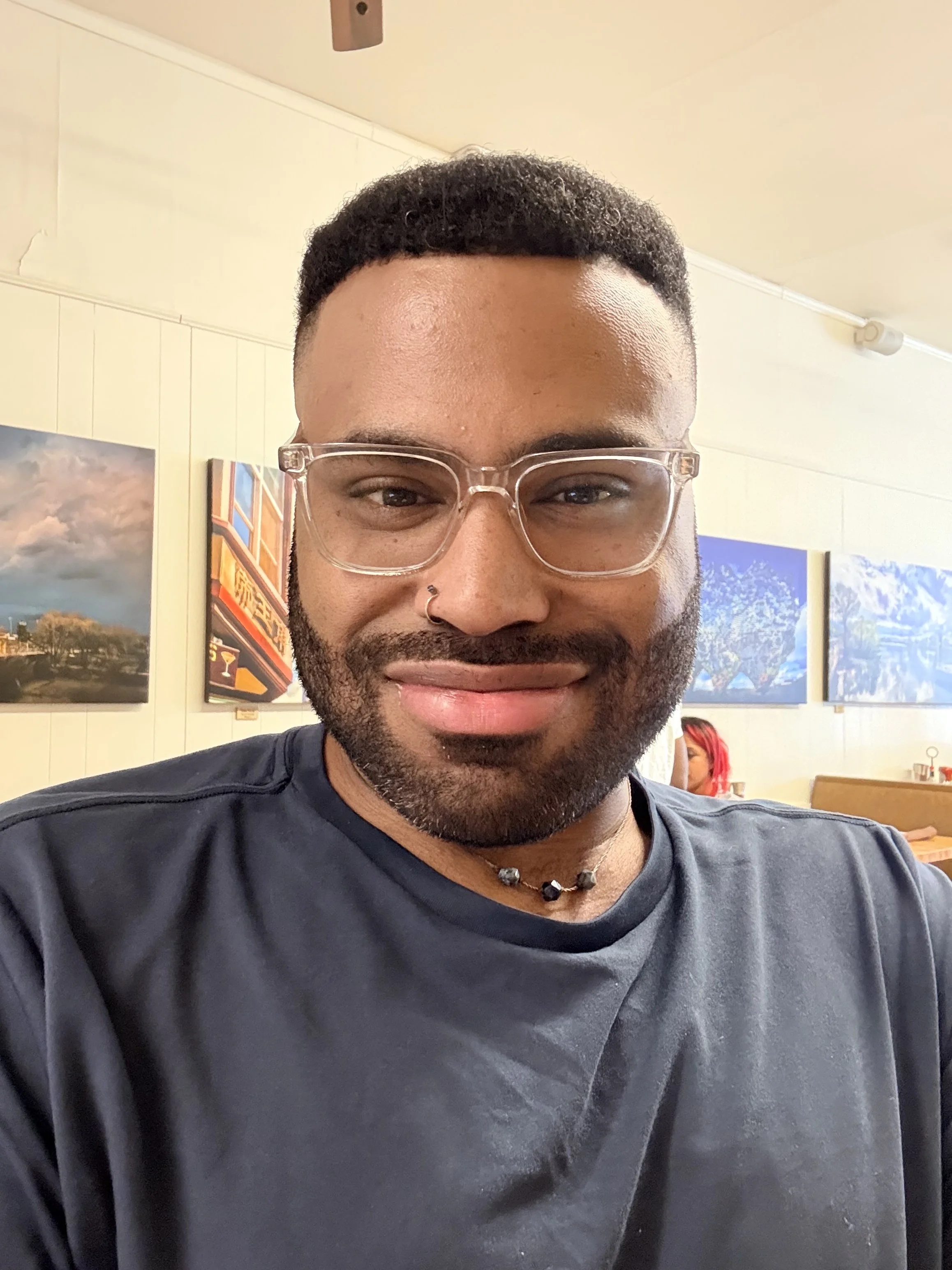 A man smiling and wearing glasses, a nose ring, a necklace, and a black t-shirt inside an art gallery with landscape paintings on the wall.