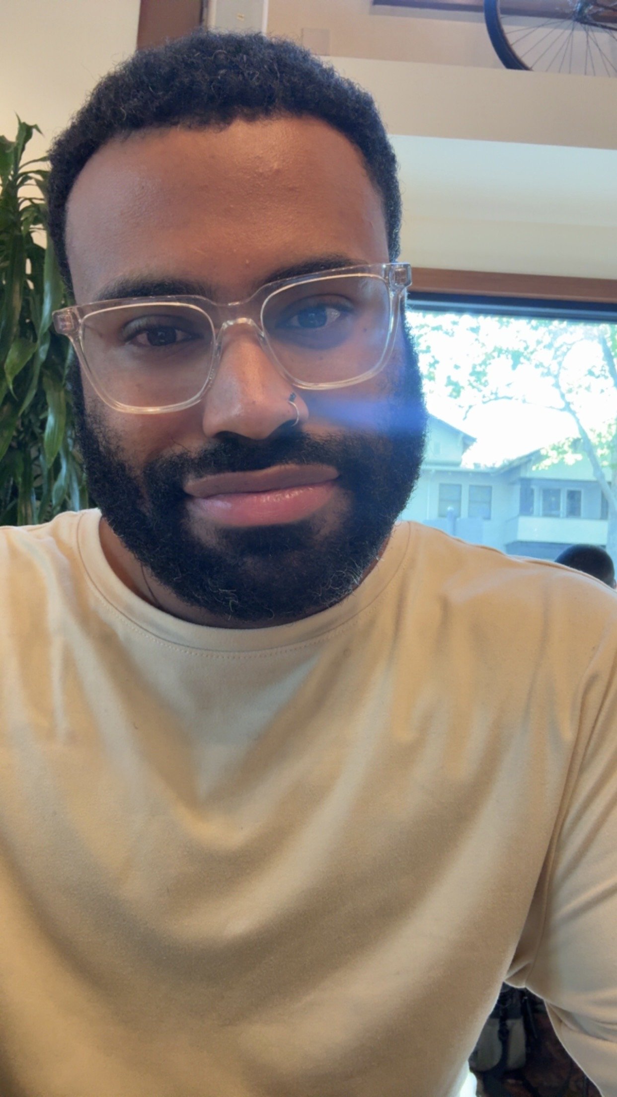 A man with a beard and glasses wearing a light-colored shirt, sitting indoors near a window with trees visible outside.