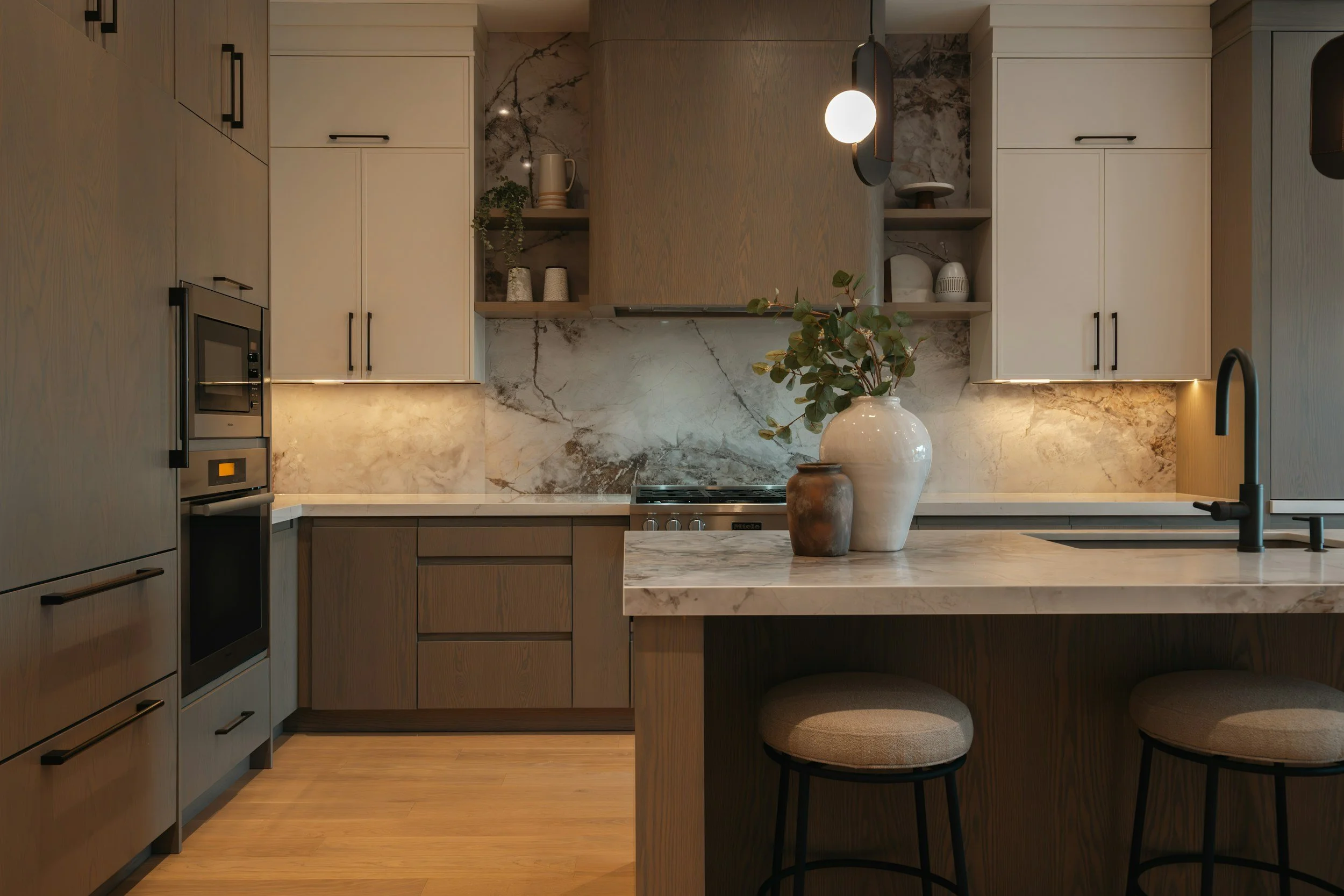 Modern kitchen with beige and brown cabinetry, marble backsplash, and a marble countertop island decorated with vases and greenery.