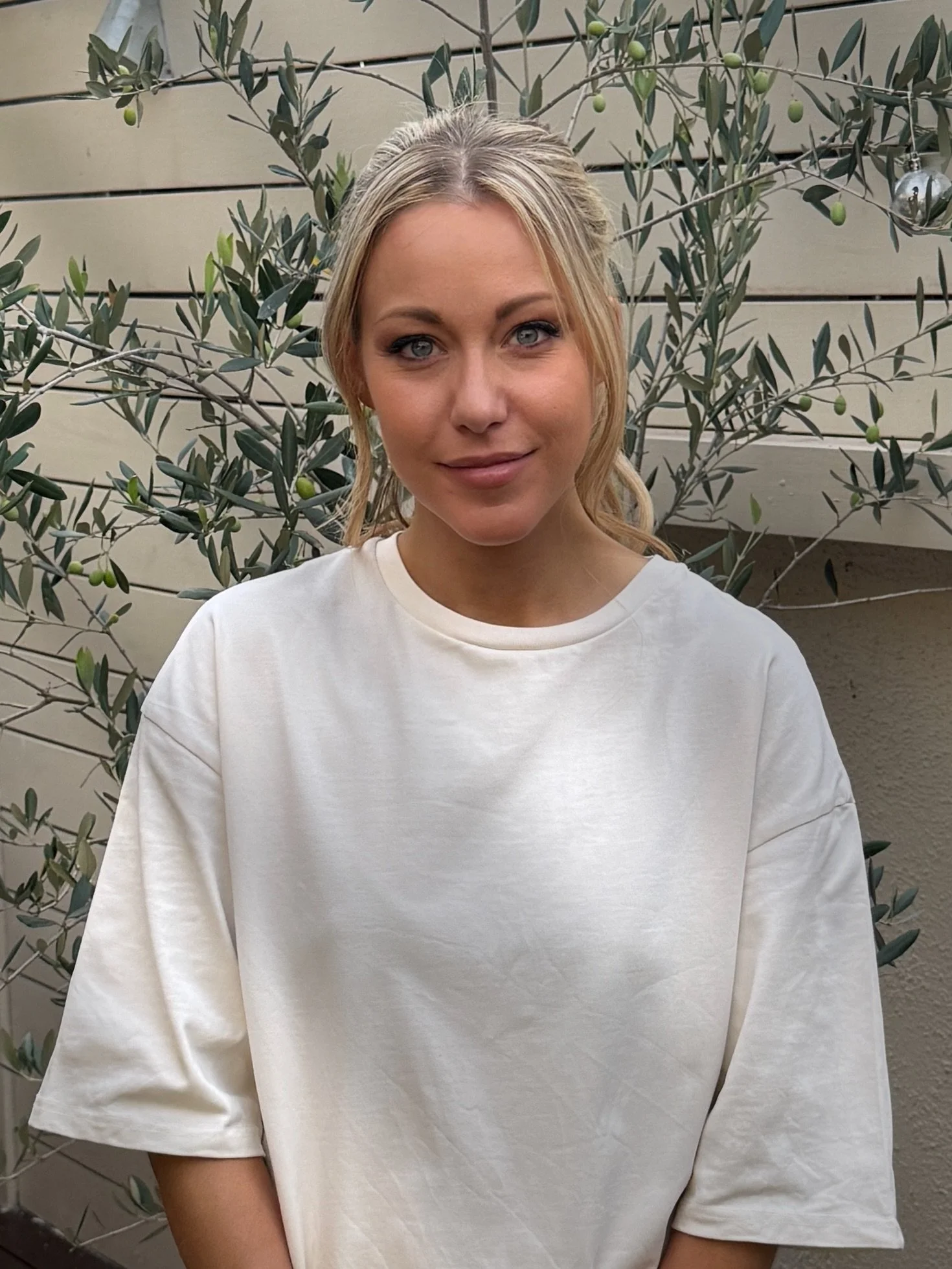 A young woman with blond hair and blue eyes standing outdoors in front of a leafy shrub with small green fruit, in front of a beige wooden fence.
