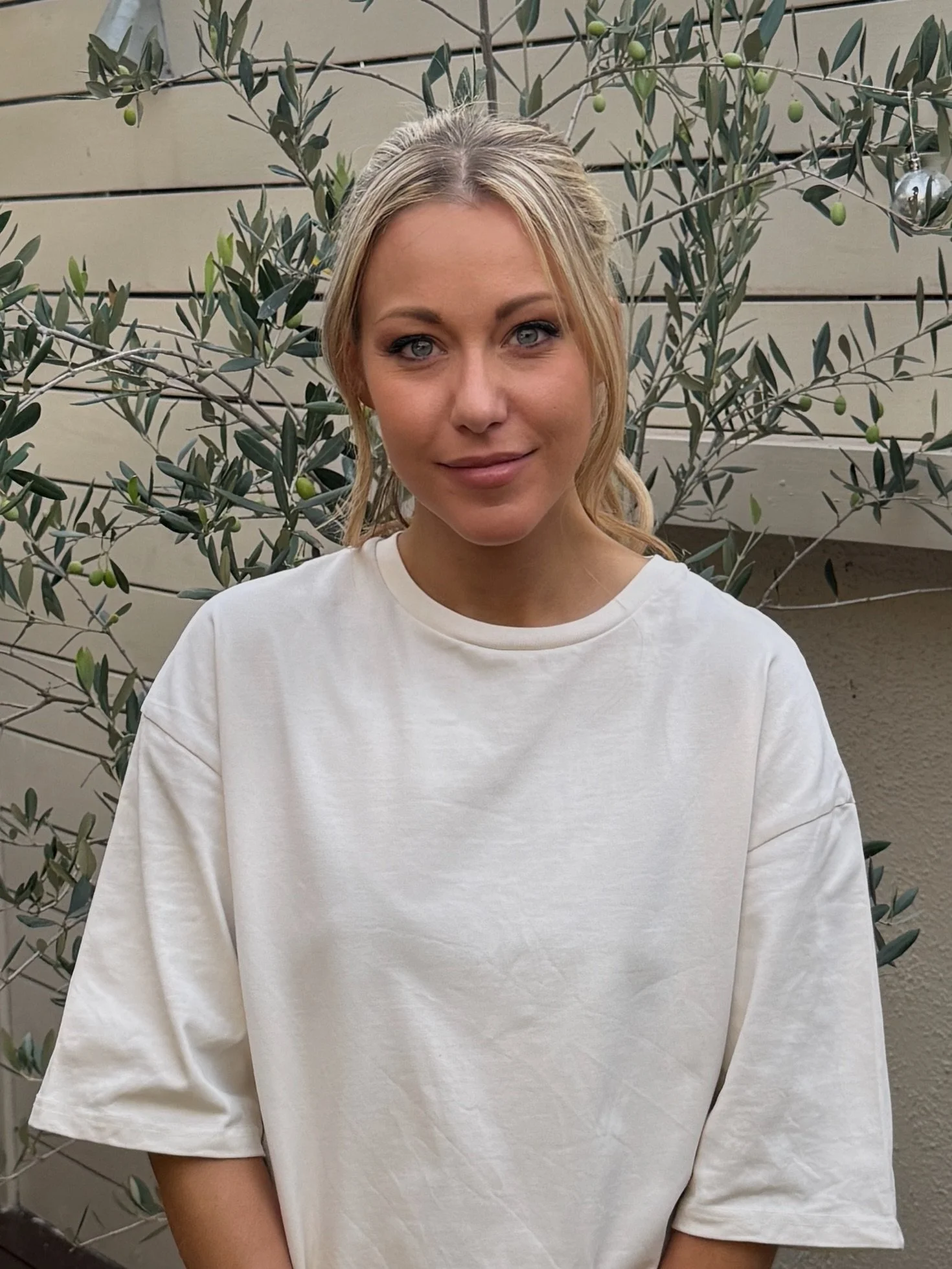 A young woman with blonde hair tied back, wearing a loose white T-shirt, standing outdoors in front of a plant with green leaves and small green and silver ornaments hanging from its branches.