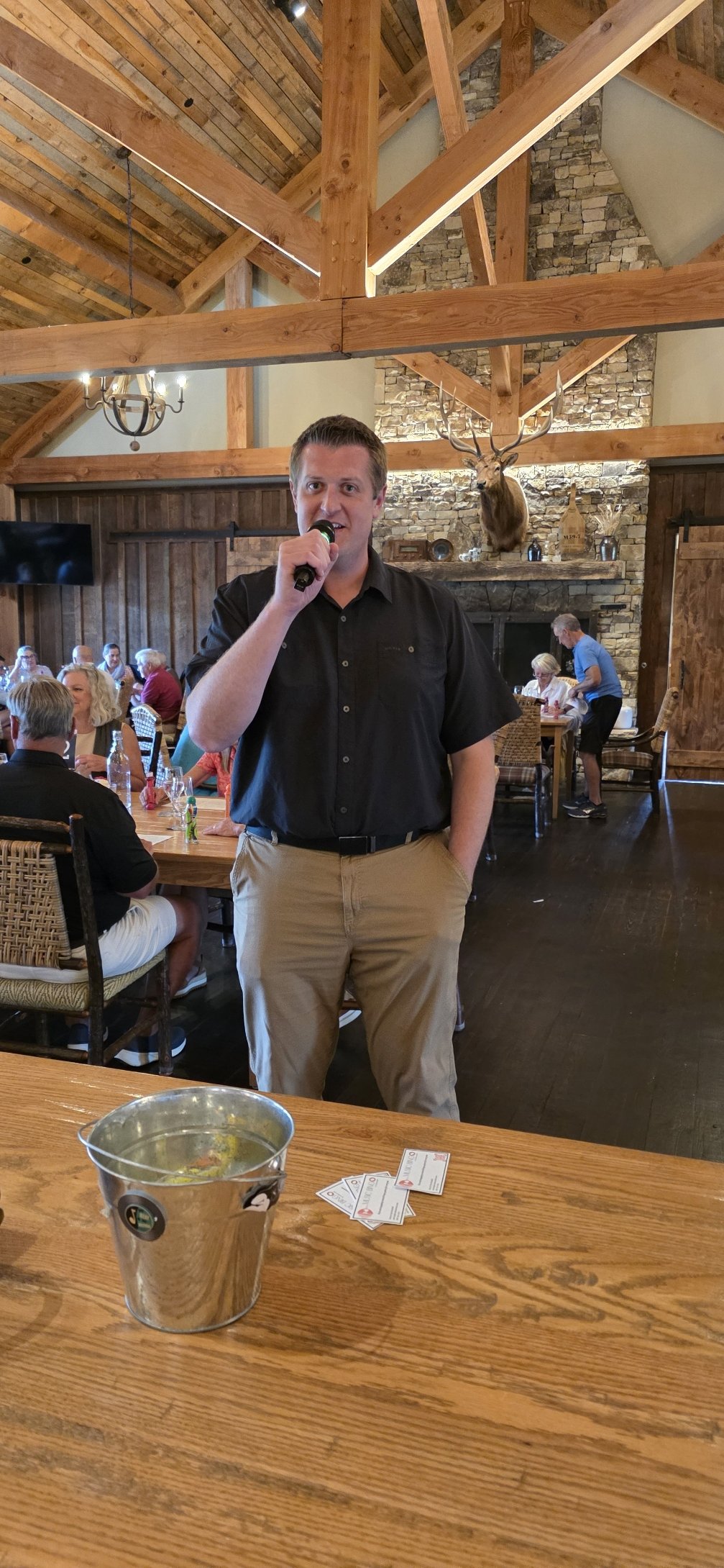 A restaurant interior with a man holding a microphone, people seated at tables, a stone fireplace, and a mounted deer head on the wall.