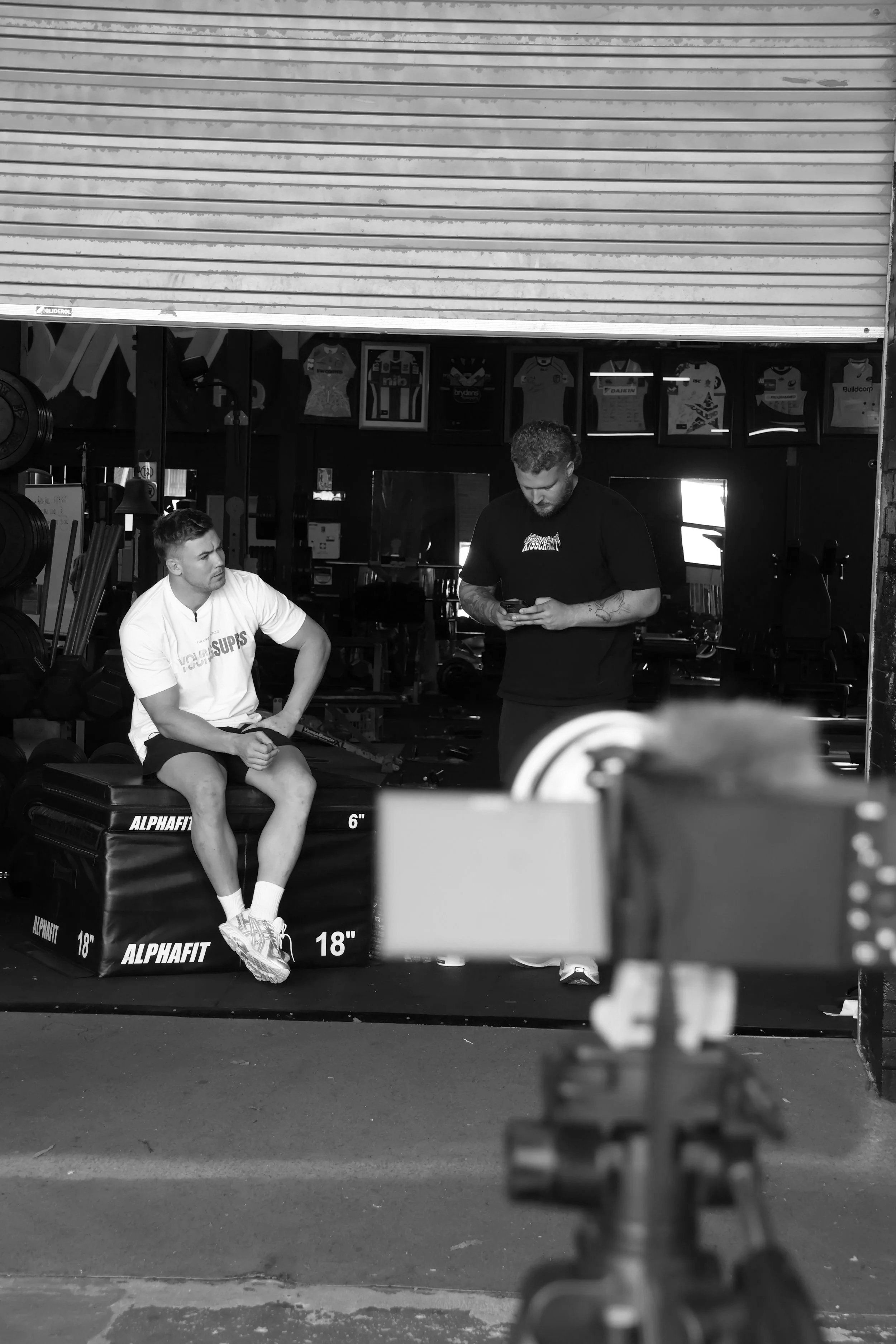 Two men in a gym. One sitting on a black plyometric box, the other standing and looking at his phone. Several framed sports jerseys are hanging on the wall behind them. A camera and tripod are in the foreground.