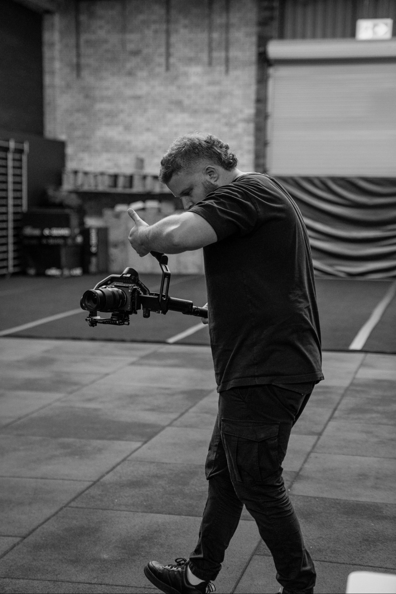 A man with curly hair and a beard holding a camera stabilizer in a large indoor space with brick walls and gym equipment