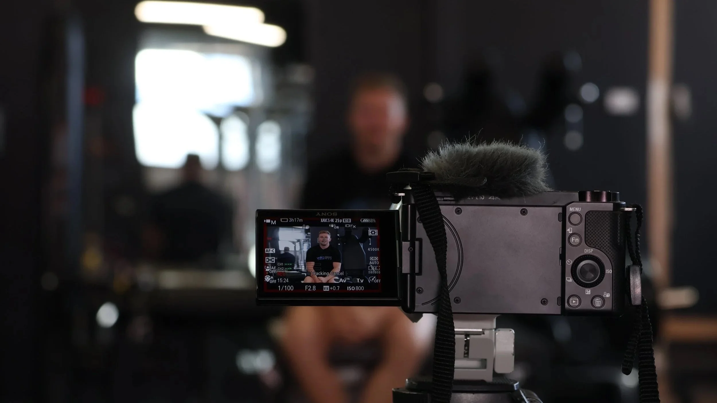 A digital camera is set up to film a person with a blurred background. The camera's display screen shows the person smiling, wearing a black shirt, with various camera settings visible.