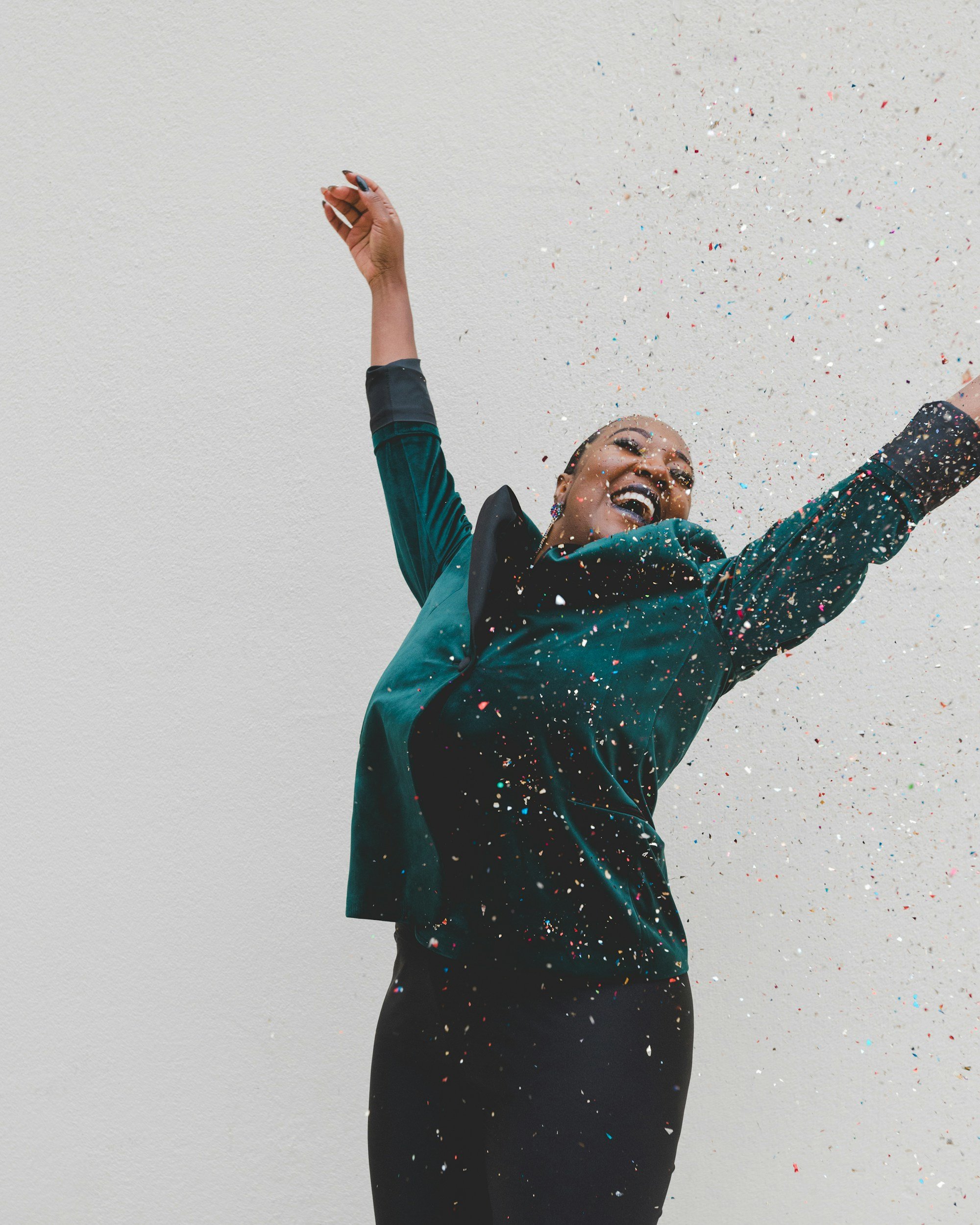 A person in a teal and black jacket laughing and throwing colorful confetti into the air against a plain white wall.