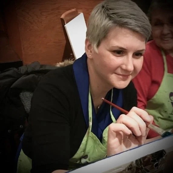 Beka Owens, a woman with short gray hair wearing a blue shirt and green apron, painting at a table in a restaurant.
