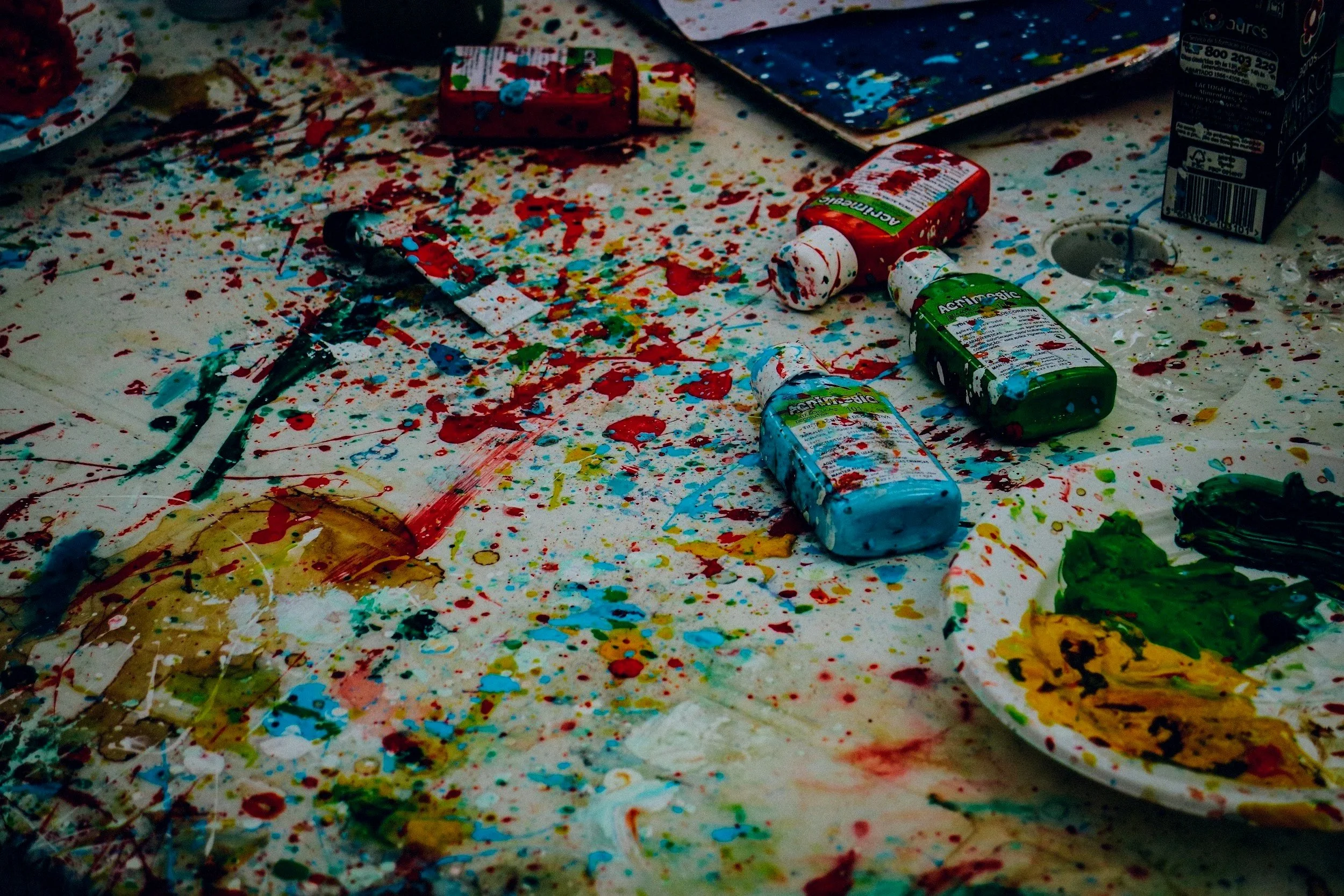 A table covered in spilled paint and art supplies, including bottles of paint, an artist's palette with green and yellow paint, and scattered paint splatters.