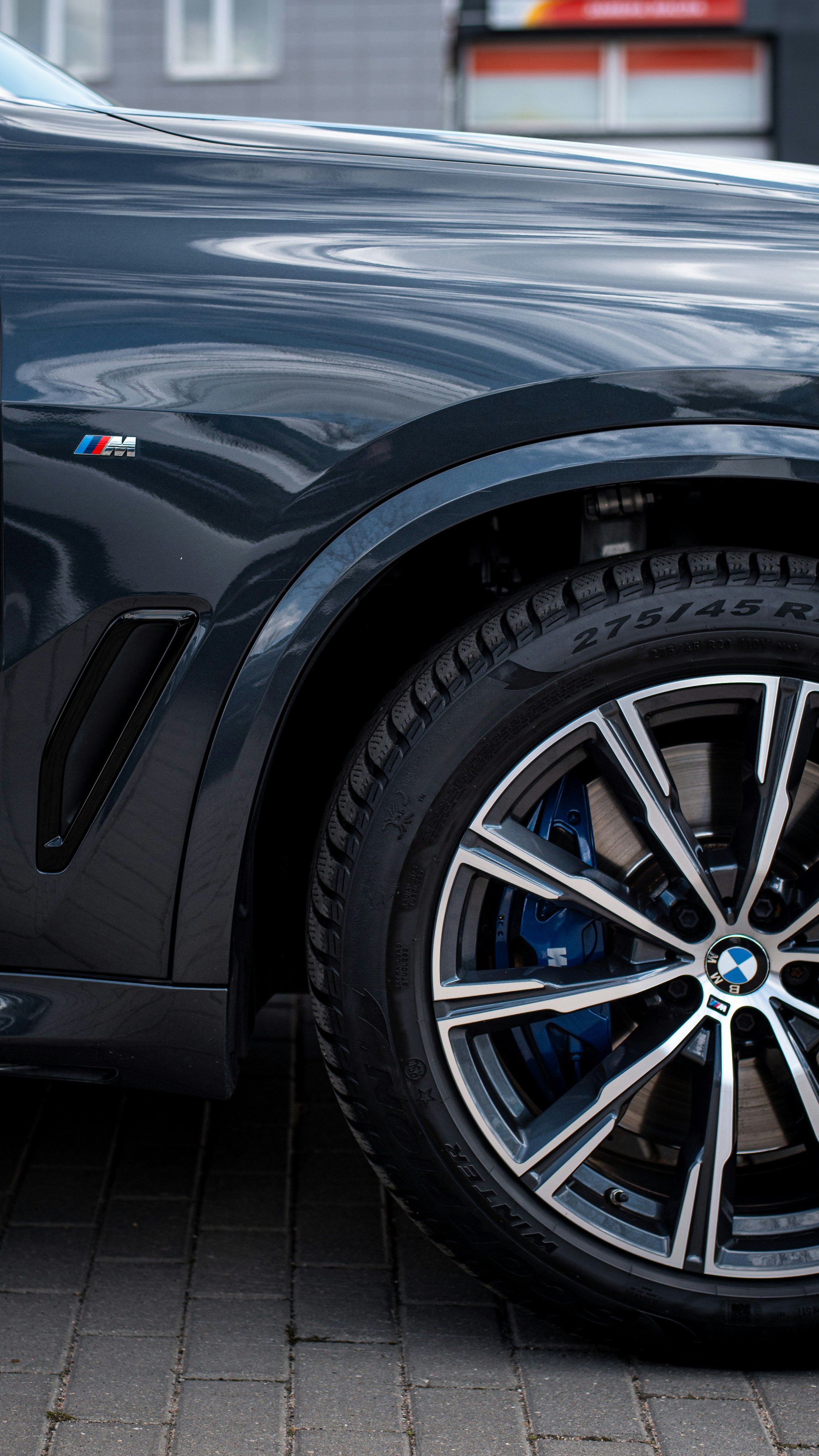 Close-up of a BMW car wheel with a blue brake caliper, tire, and part of the dark gray body of the vehicle, with a BMW emblem and M badge visible.