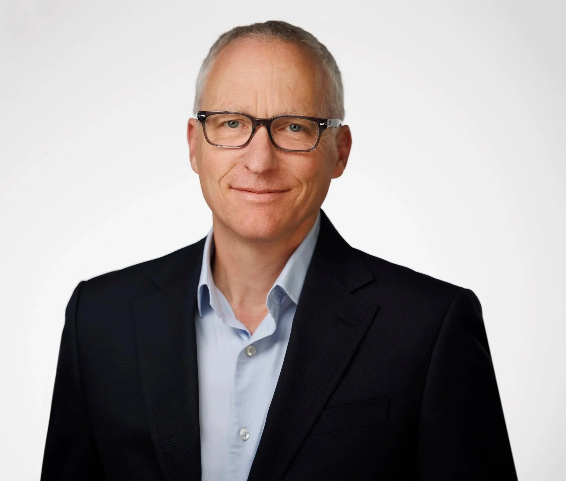 A middle-aged man with short gray hair and blue eyes smiling, wearing black glasses, a dark suit jacket, and a light blue collared shirt against a plain white background.