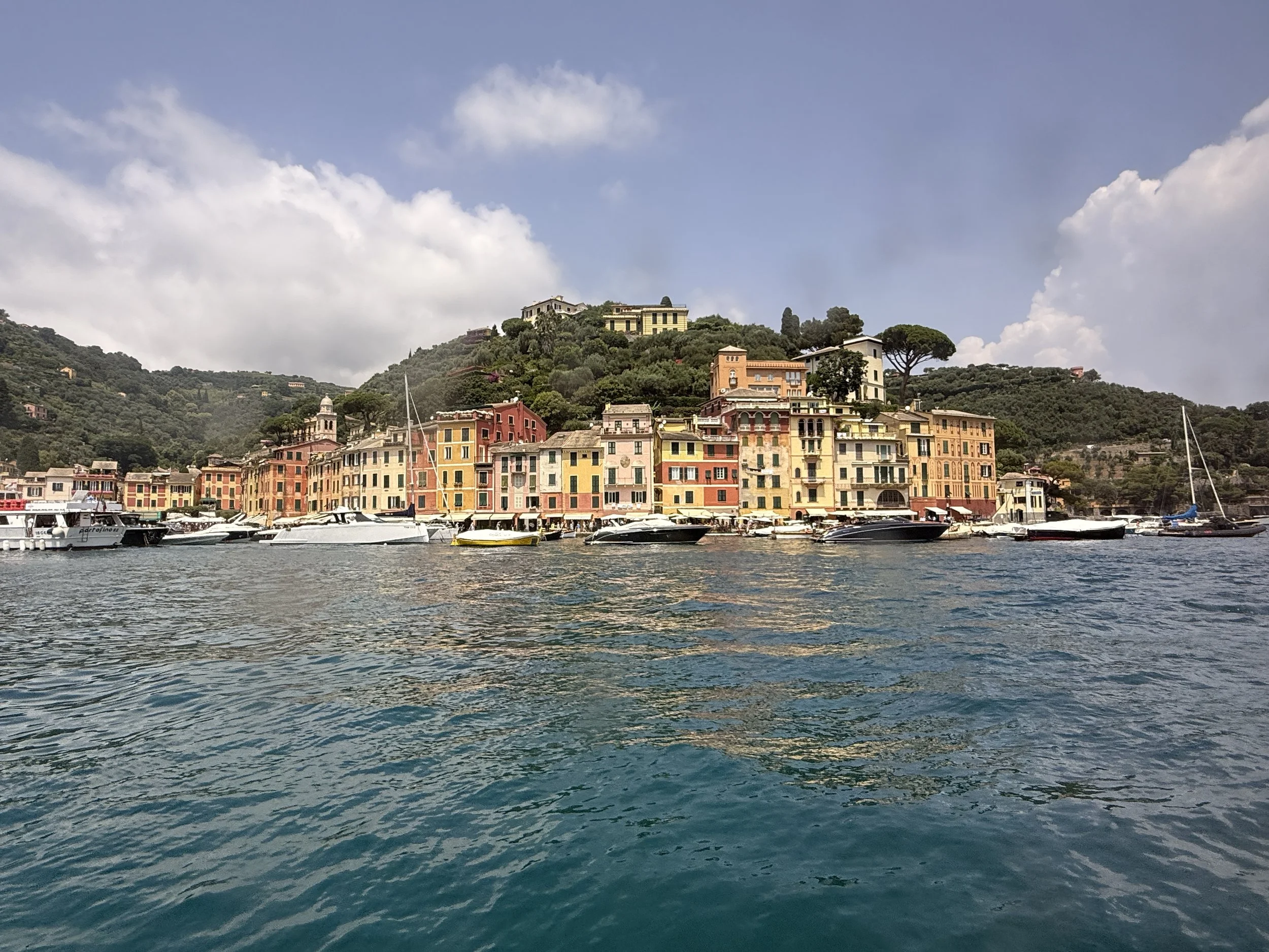 The colorful buildings of Portofino lining the waterfront in a coastal town with boats docked in the water and lush green hills in the background under a partly cloudy sky.