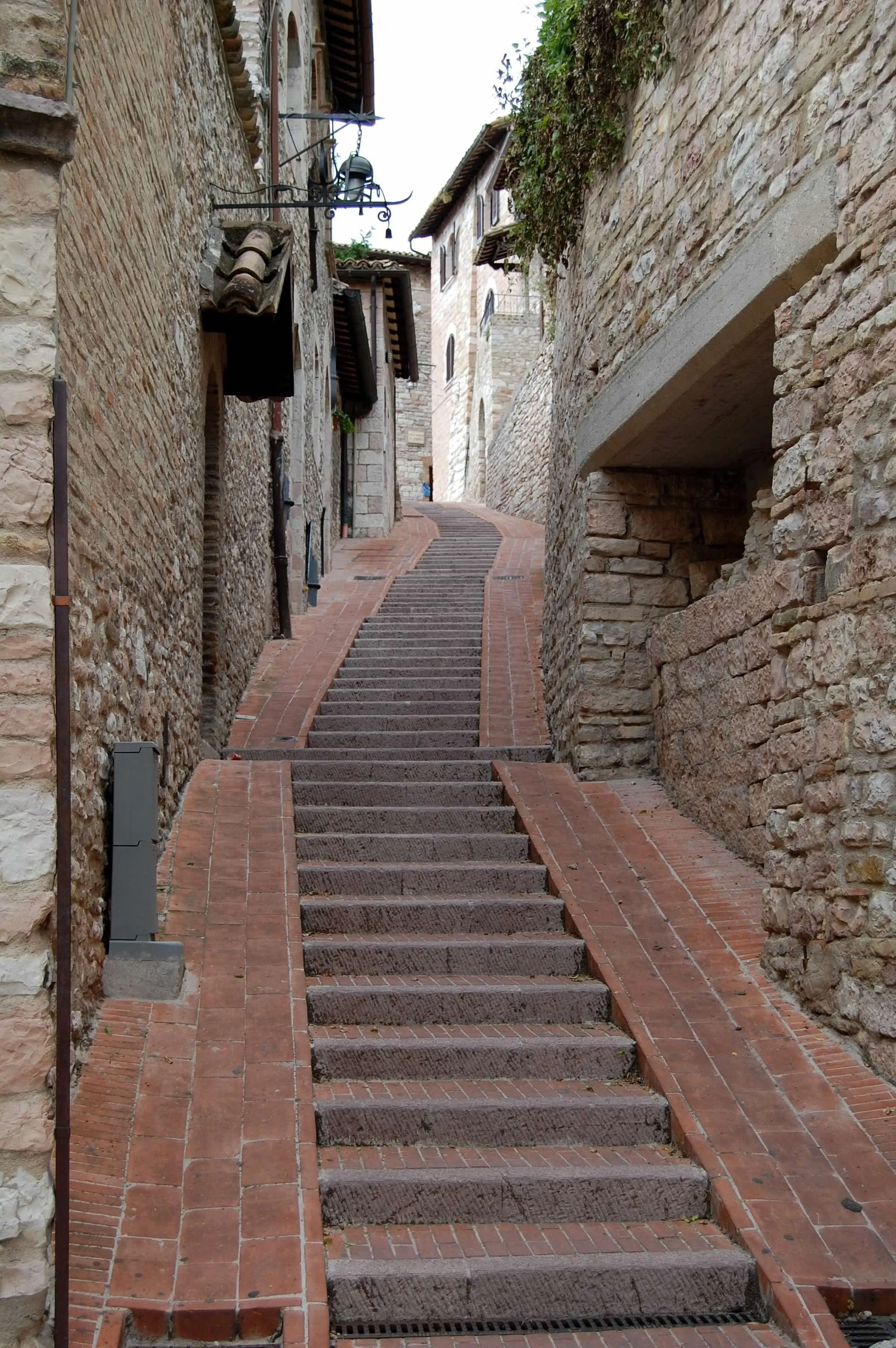 A narrow, sloped alleyway with stone stairs and red brick borders, flanked by old stone buildings with tiled roofs in a European village.