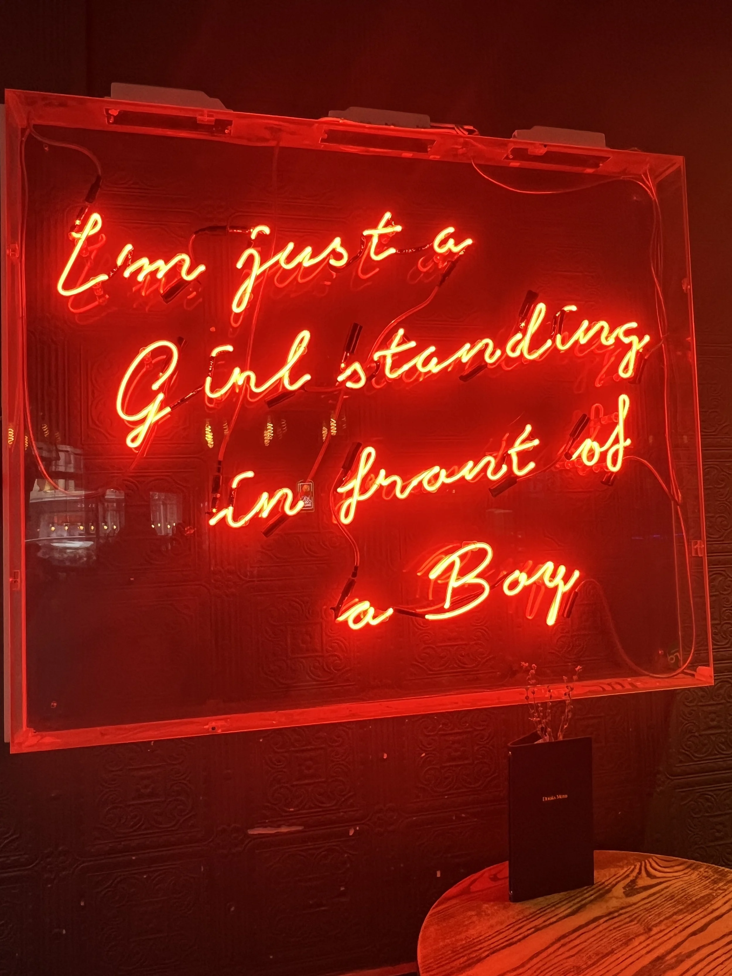 Neon sign reading 'I'm just a girl standing in front of a boy' in red cursive on a dark wall.