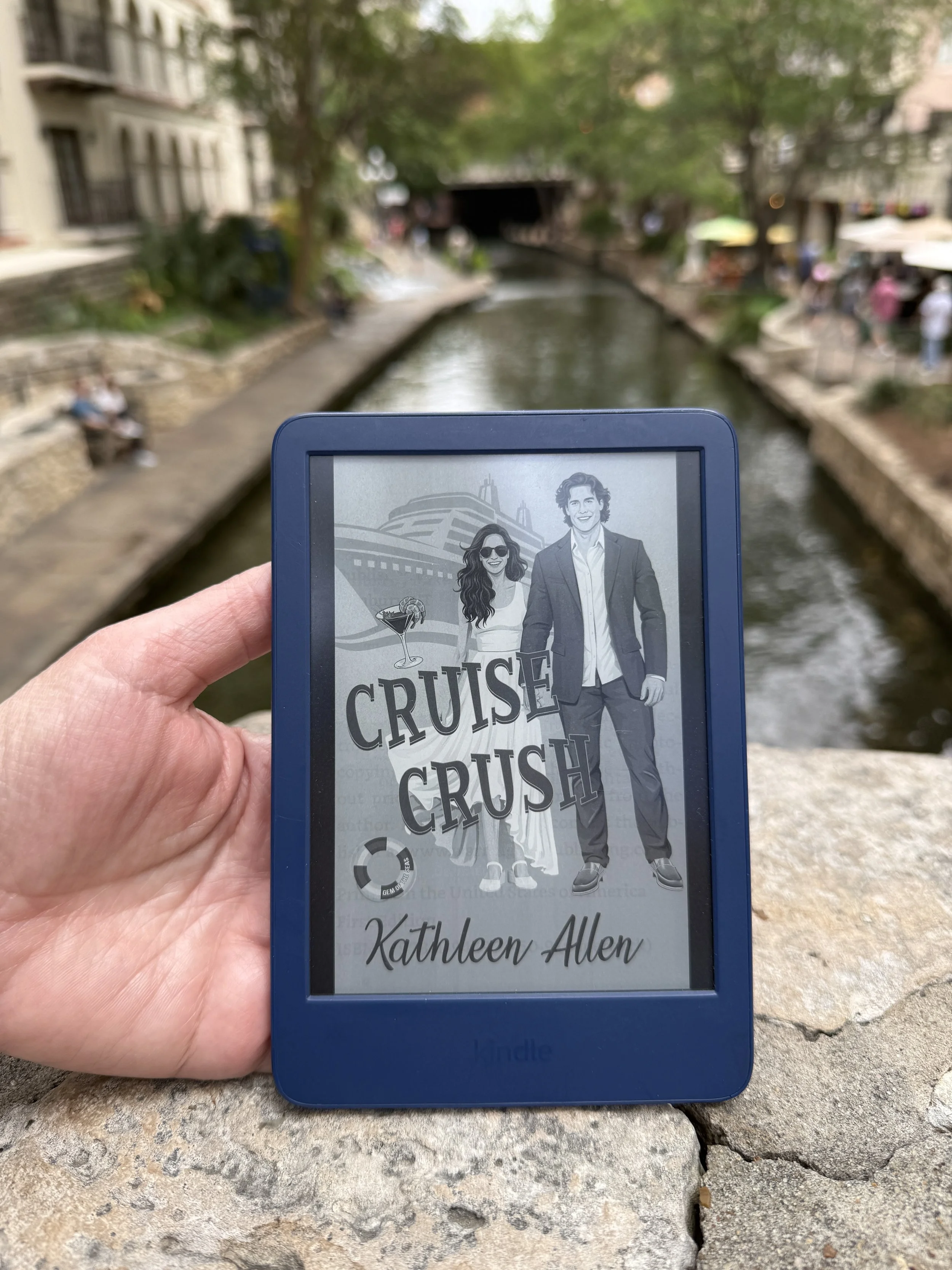 E-reader displaying the book cover of 'Cruise Crush' by Kathleen Allen, held in front of a city canal with a bridge, trees, and people walking along the water.
