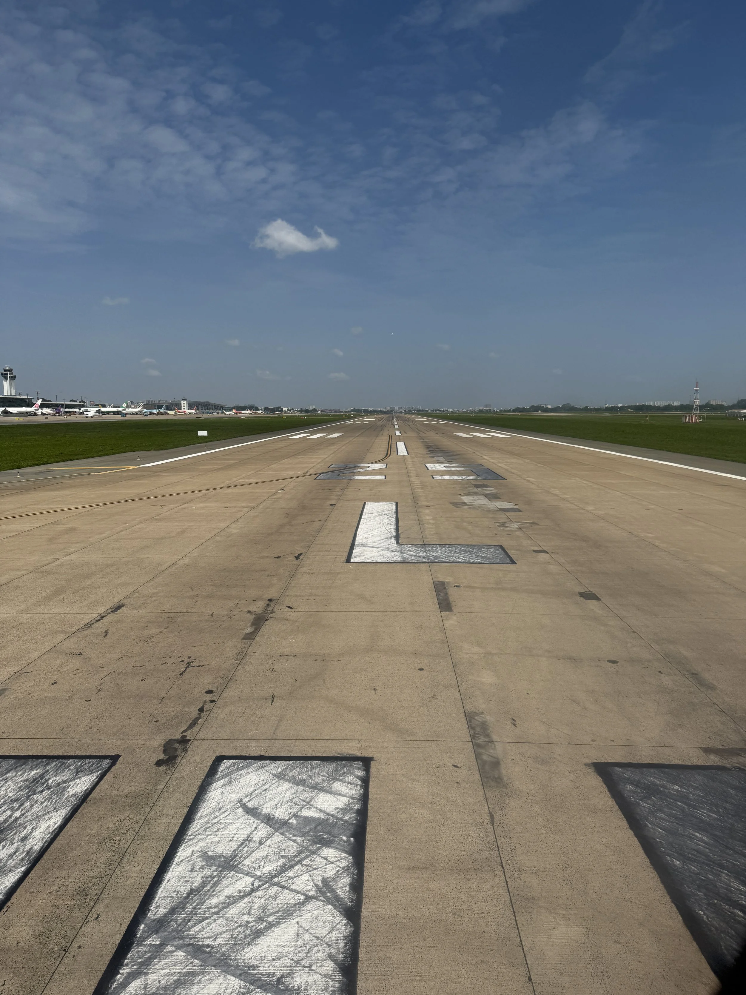 View of an airport runway with markings, stretching into the distance under a blue sky with scattered clouds, and airport buildings visible on the horizon.