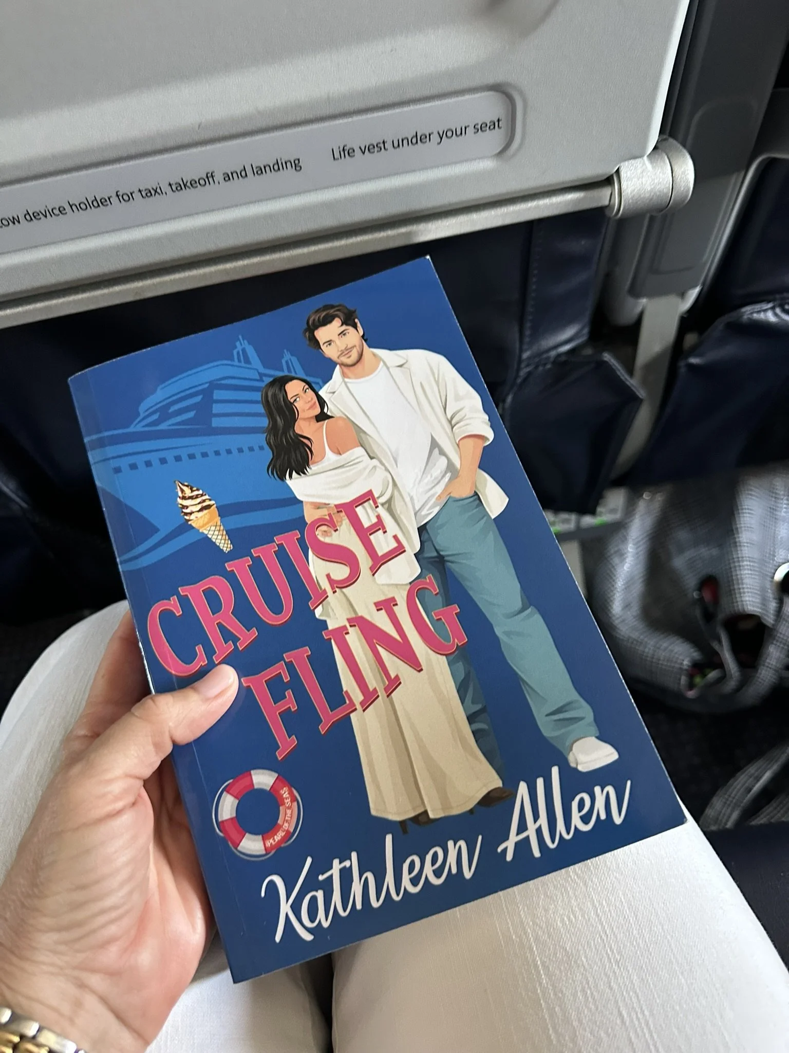 Holding a paperback copy of Cruise Fling by Kathleen Allen on an airplane, part of the Pinnacle Cruise Line series — the perfect travel companion for a flight, vacation, or beach read.