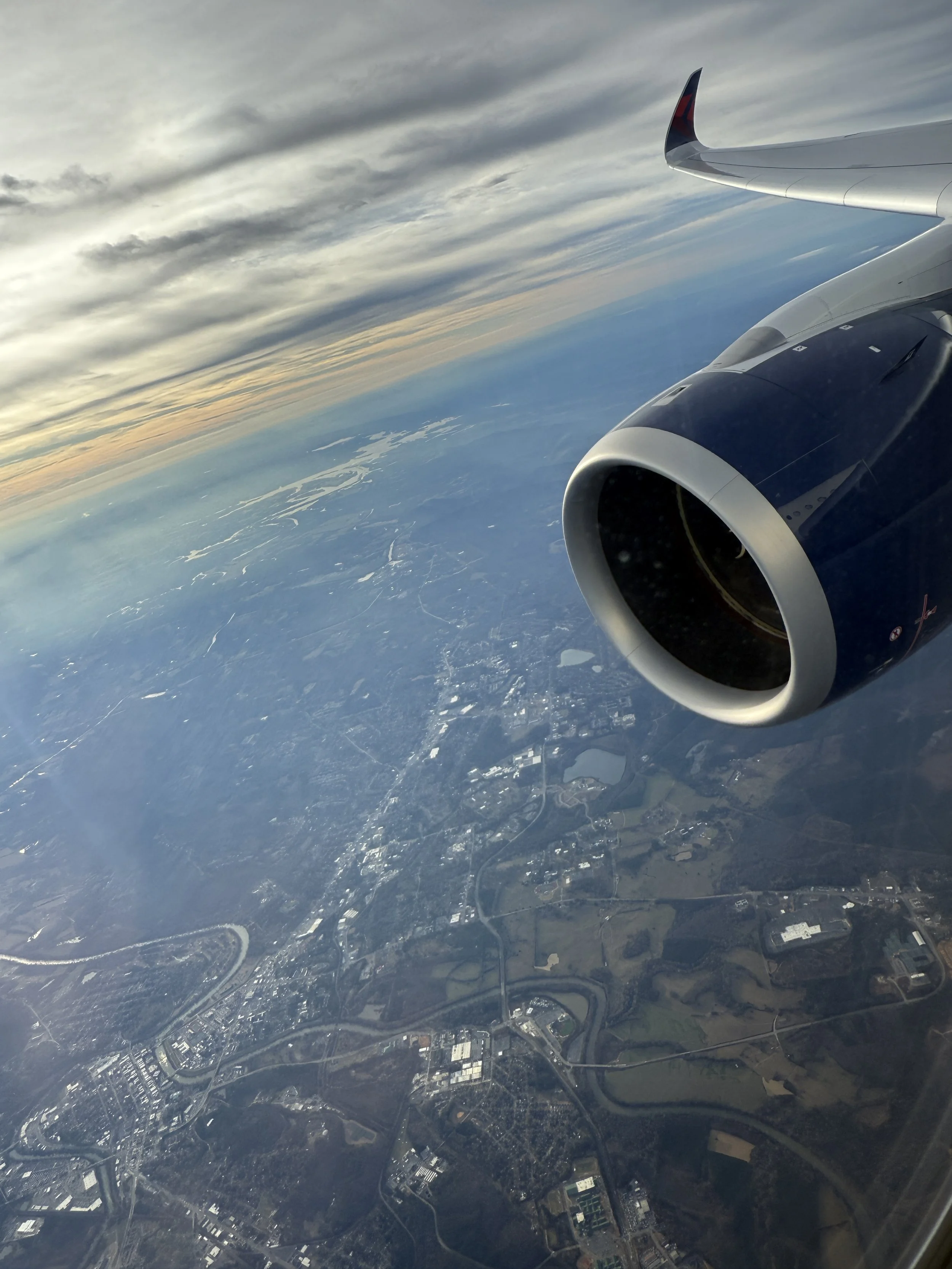 View from an airplane window showing the engine, wingtip, and the landscape below with clouds, river, and cityscape during sunset.
