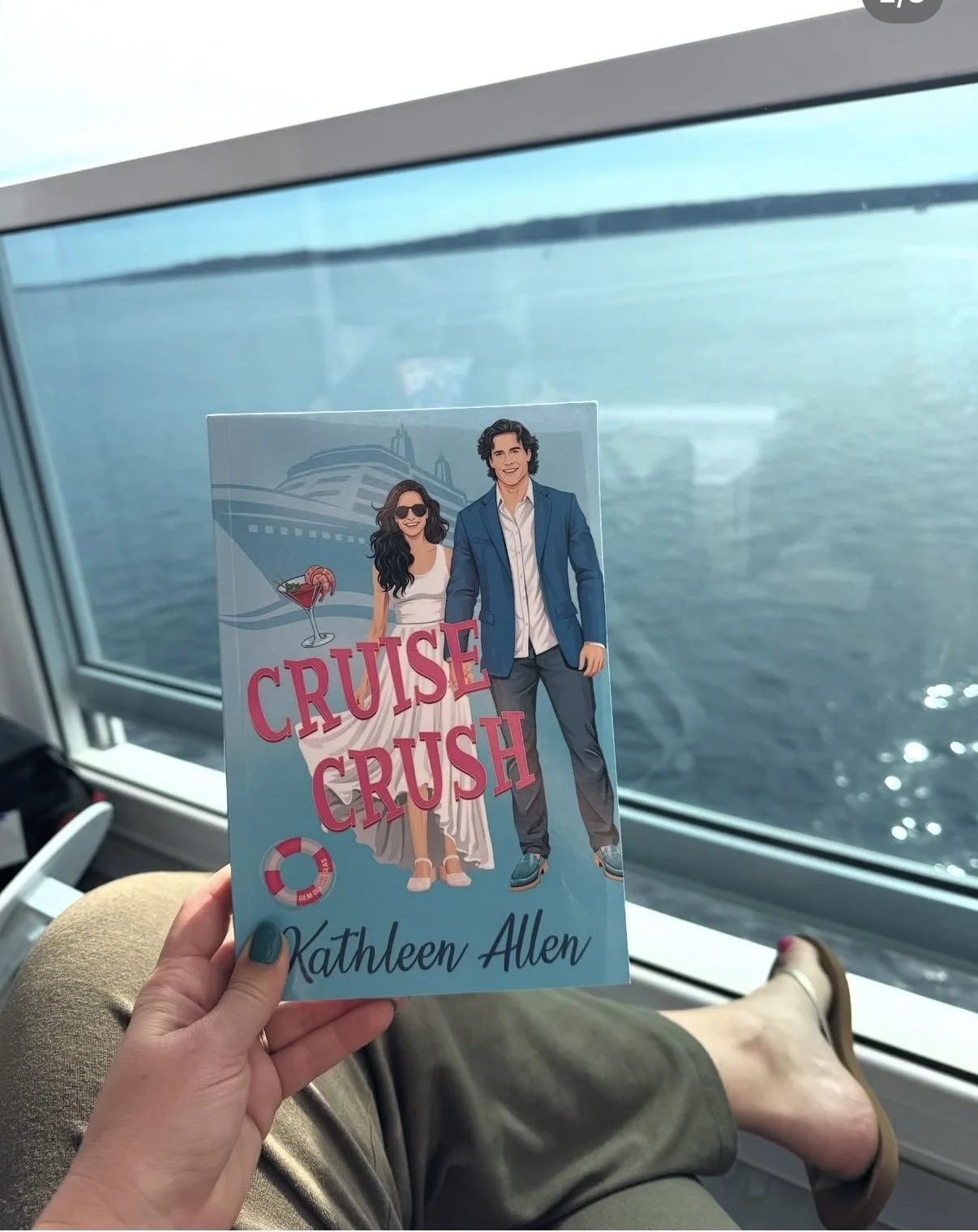 A hand holding a book titled 'Cruise Crush' by Kathleen Allen, with an illustration of a couple on a cruise, a cruise ship in the background, and a cocktail glass with a pink drink. Someone's legs and feet are visible, and they are seated near a window showing water outside.