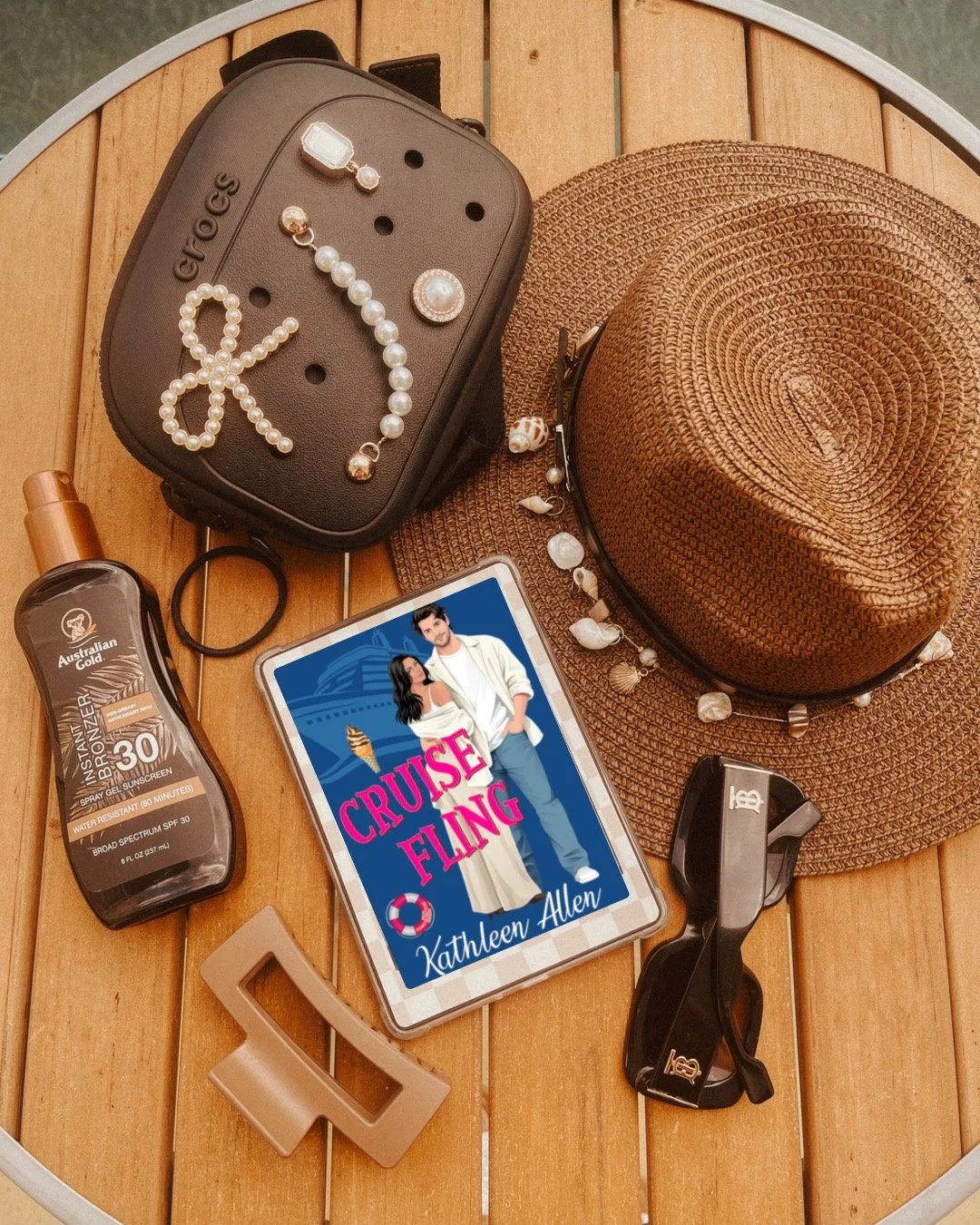 A wooden table with a straw hat, sunglasses, a book titled "Cruise Fling" by Kathleen Allen, a tube of Australian Gold sunscreen, a small black bag with jewelry including pearl earrings, a pearl bracelet, and a pearl hair clip, a gel hair tie, and a beige decorative letter T.