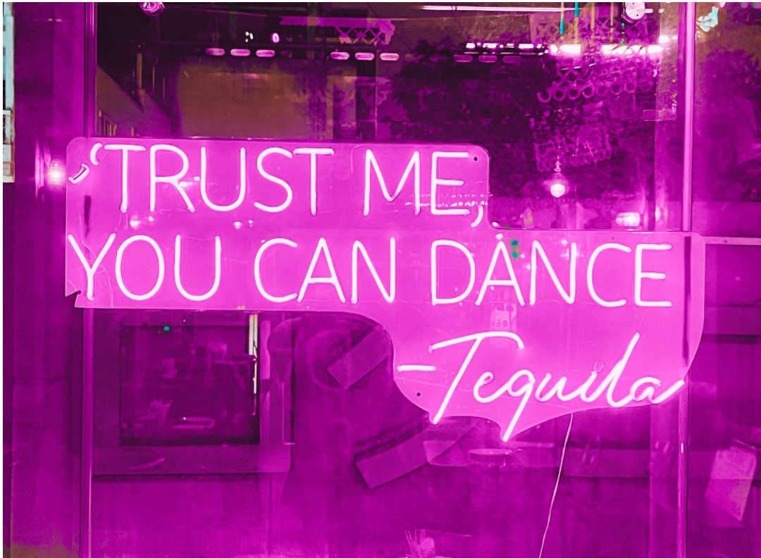 Pink neon sign with the quote 'Trust me, you can dance - Tequila' in a bar or nightclub setting.