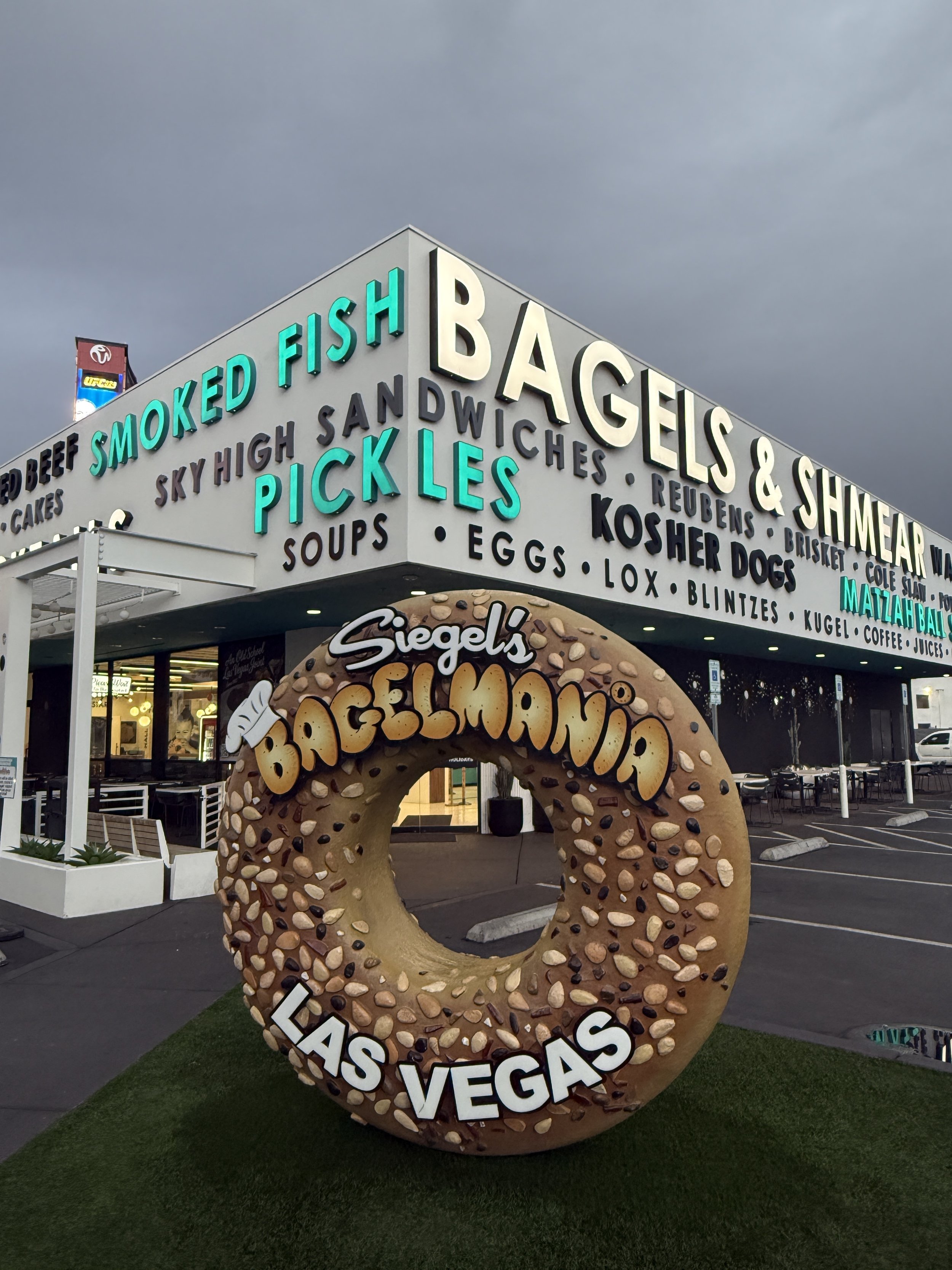 Exterior of Siegel's Bagelmania in Las Vegas, with a giant bagel sign at the entrance and various neon signs above the building advertising smoked fish, bagels, schmears, and other deli items.