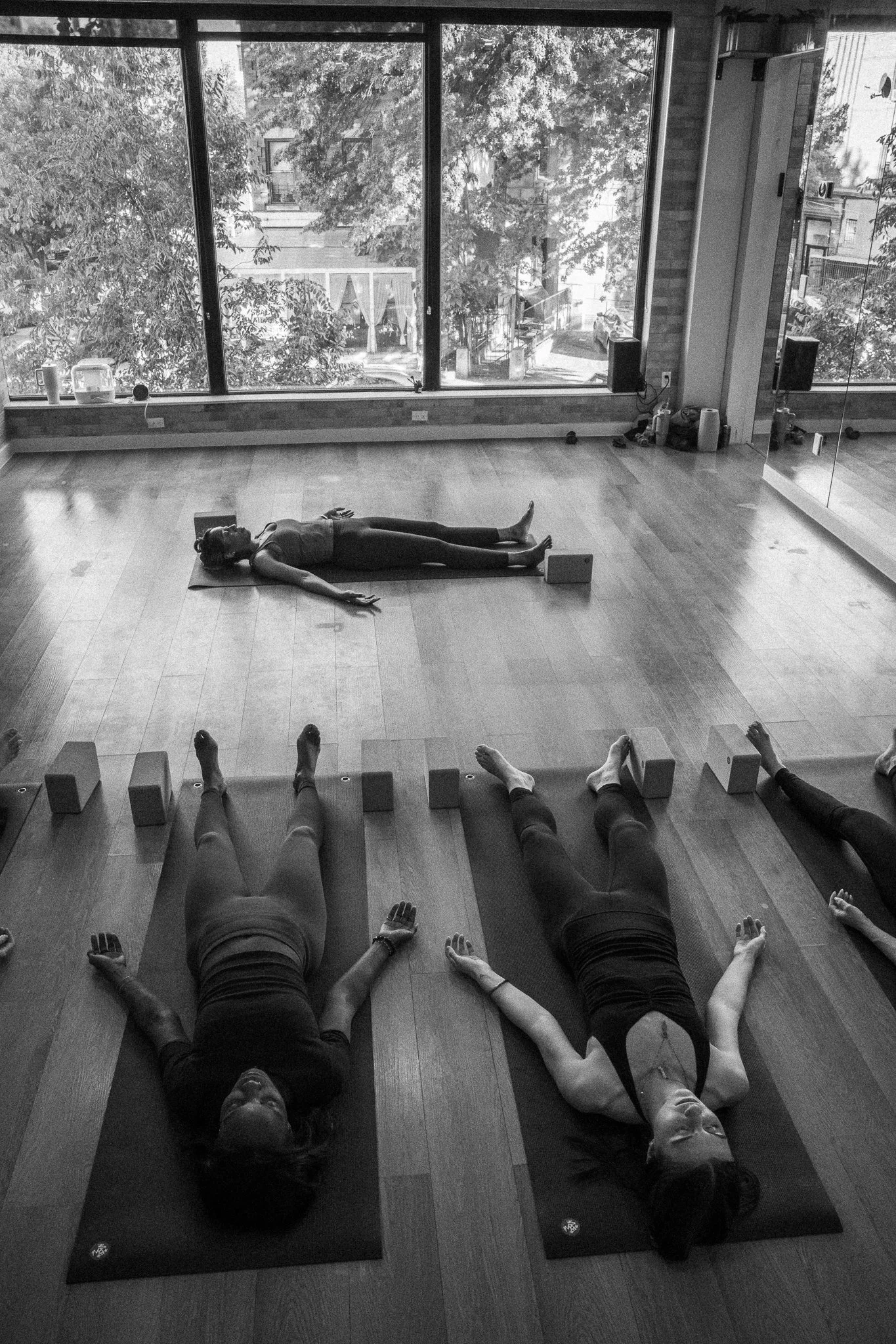 People participating in a yoga class lying on mats with their arms outstretched, viewed from the front, in a room with large windows, wood flooring, and a reflection of the room in a mirror.