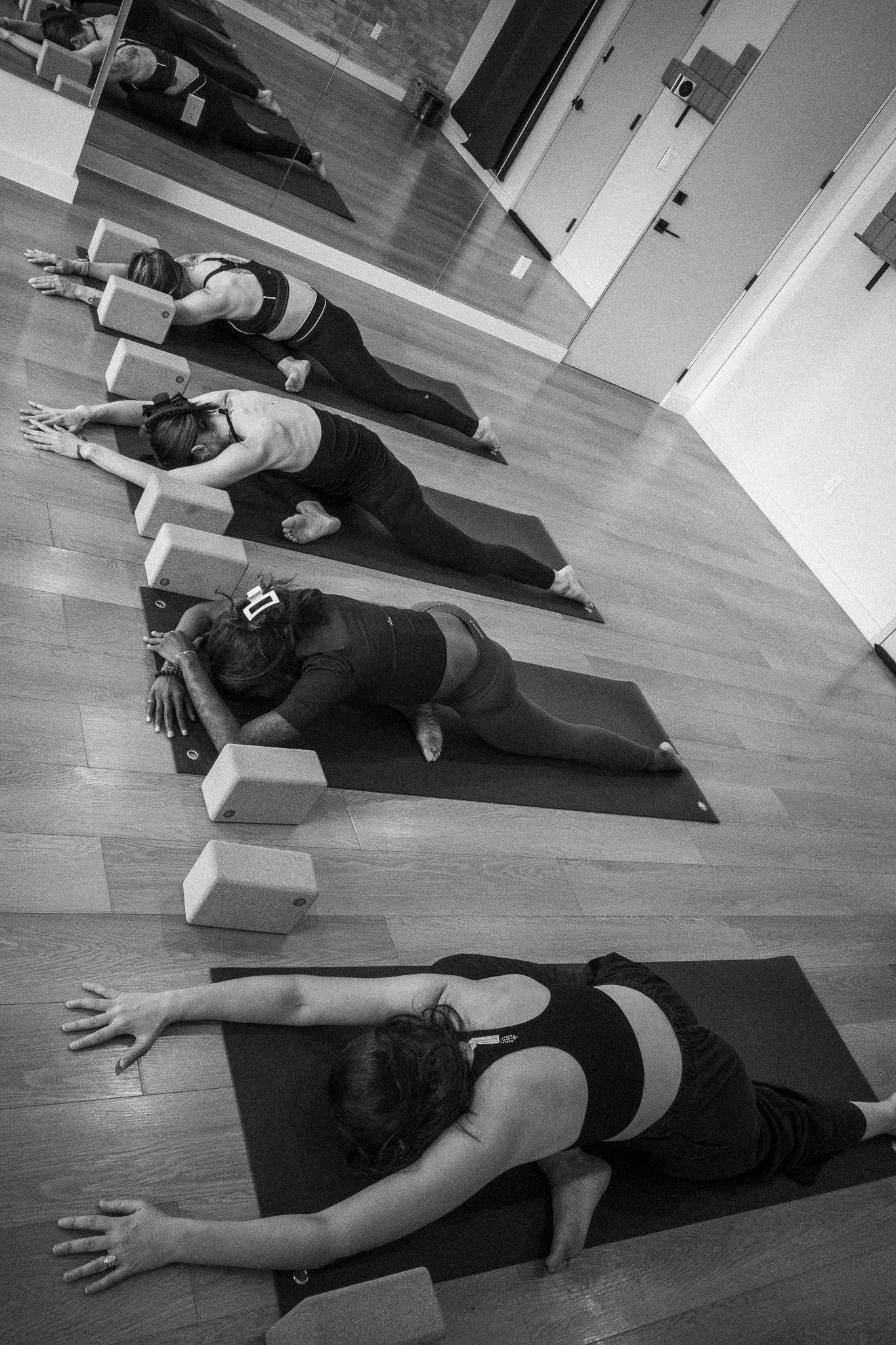 Four women participating in a yoga class, lying on black mats and stretching in a large room with a mirrored wall and wooden floor.