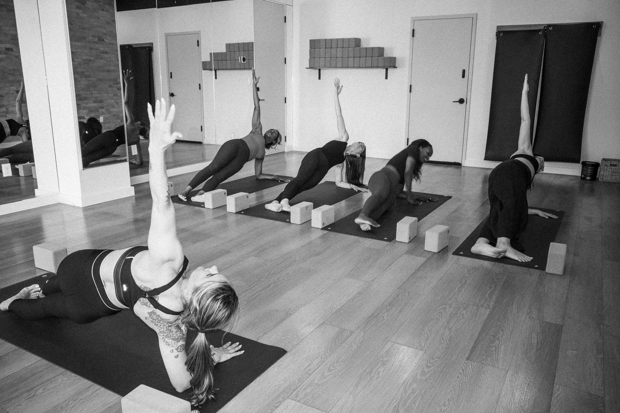 Group of women practicing yoga in a studio, some in downward dog pose, with yoga blocks and mats, a large mirror, and a brick wall in the background.
