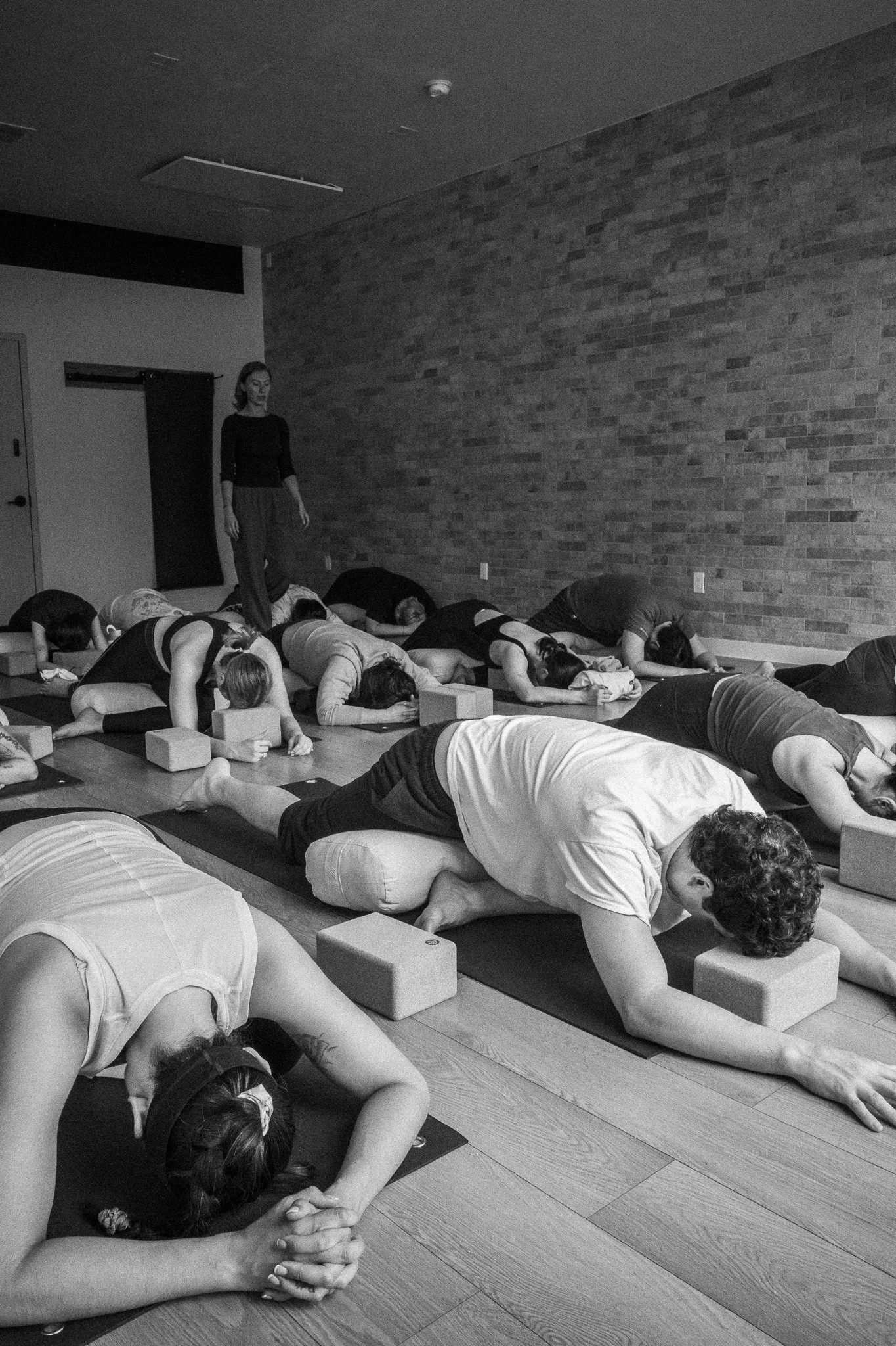 A group of people practicing yoga in a studio, performing the child pose position with their arms extended and foreheads on the mats, with a woman standing at the front of the room.