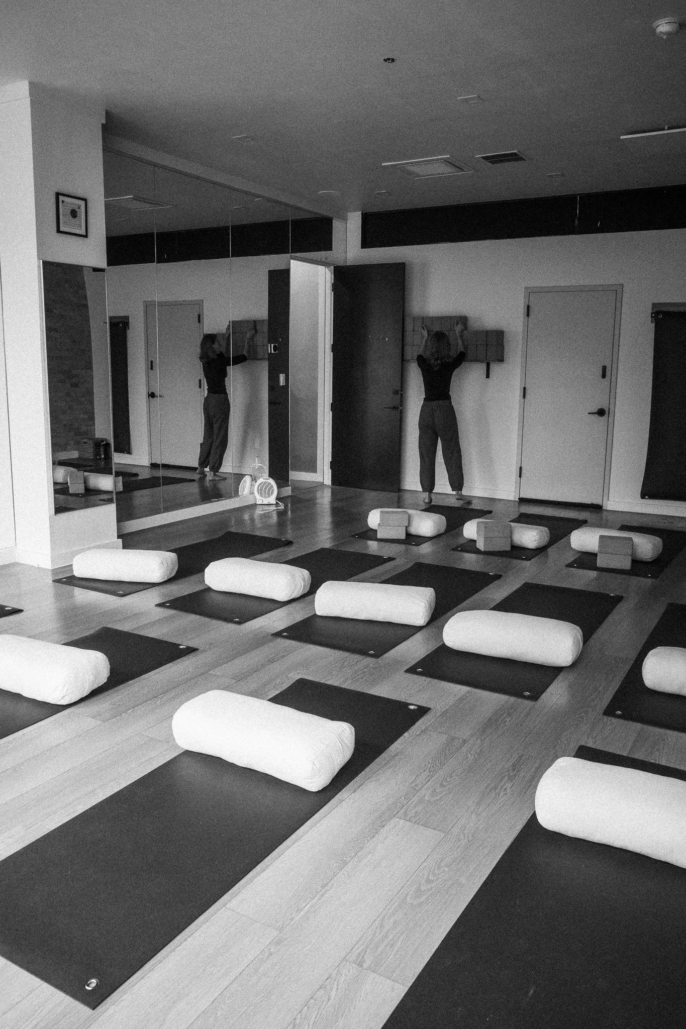 Yoga studio with black mats, bolsters, and foam blocks on wooden floor, mirrored wall on one side, woman standing and reaching towards the wall, yoga props arranged on floor.