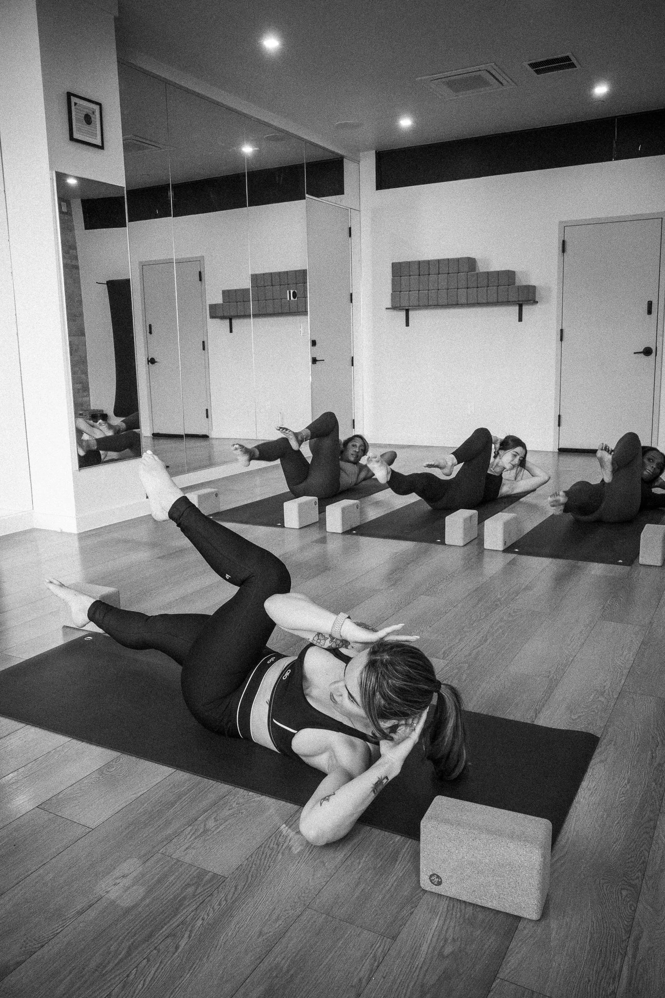 People doing yoga with legs bent and arms reaching back, lying on yoga mats in a fitness studio, with a large mirror covering the wall and shelves with small blocks.
