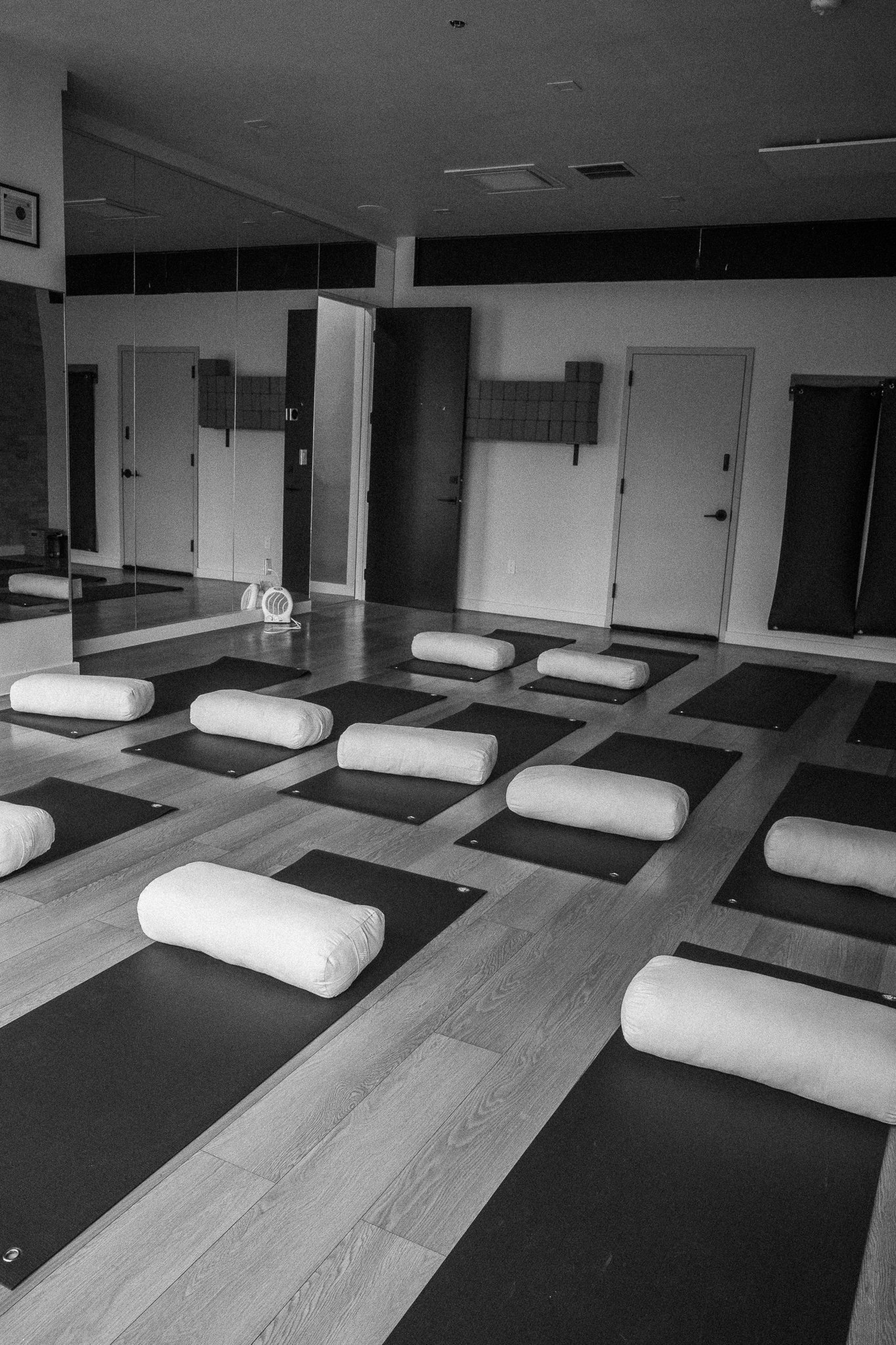 A yoga or meditation studio with yoga mats and bolsters arranged on a wooden floor, with a large mirror on the wall and minimal decor.