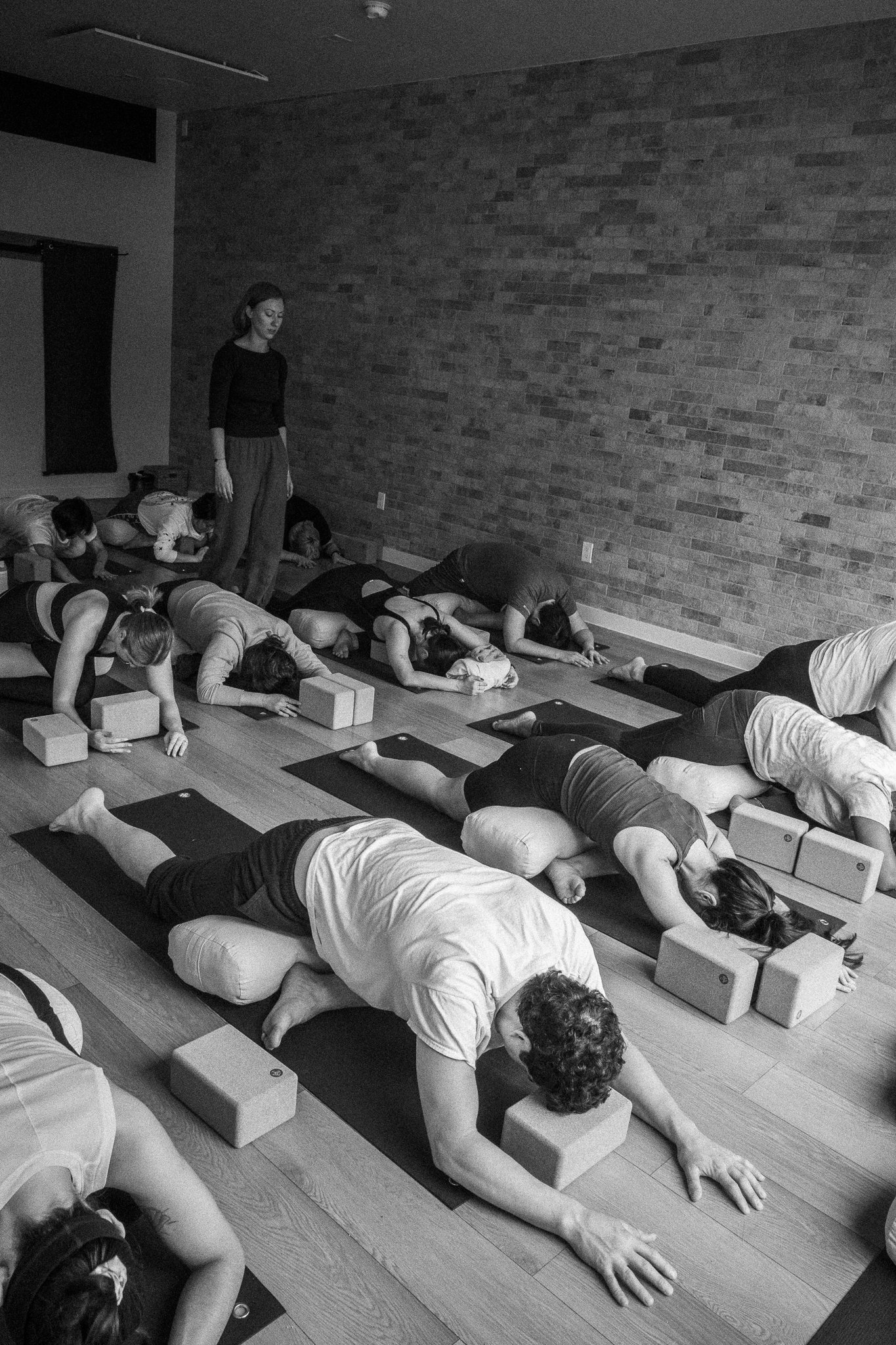 People practicing yoga in a studio with a woman standing in the middle, and they are doing downward dog poses with yoga blocks and mats.