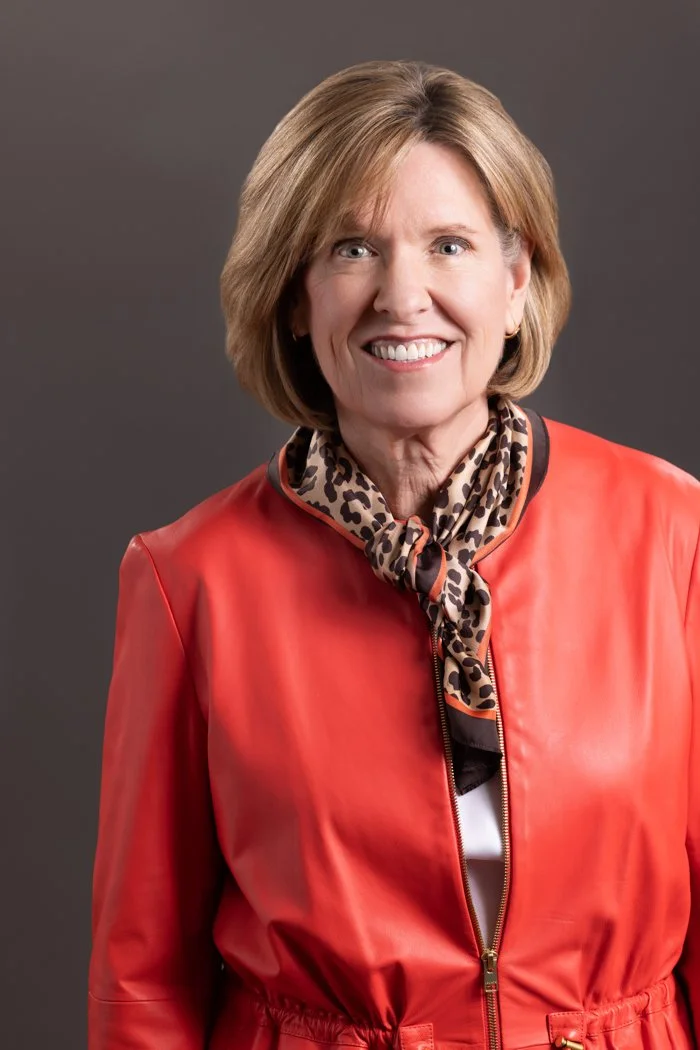 Retired Judge Catherine Hammond wearing a red jacket and a leopard print scarf against a dark brown background.