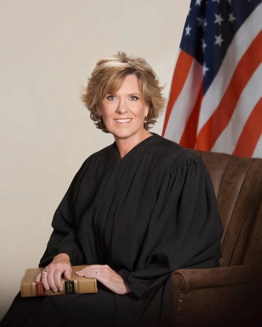 A woman wearing a black judge's robe sitting on a brown chair, holding a book, with an American flag in the background.
