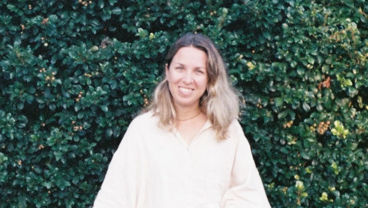 A woman with shoulder-length blonde hair smiling at the camera, standing outdoors in front of a dense green leafy hedge.