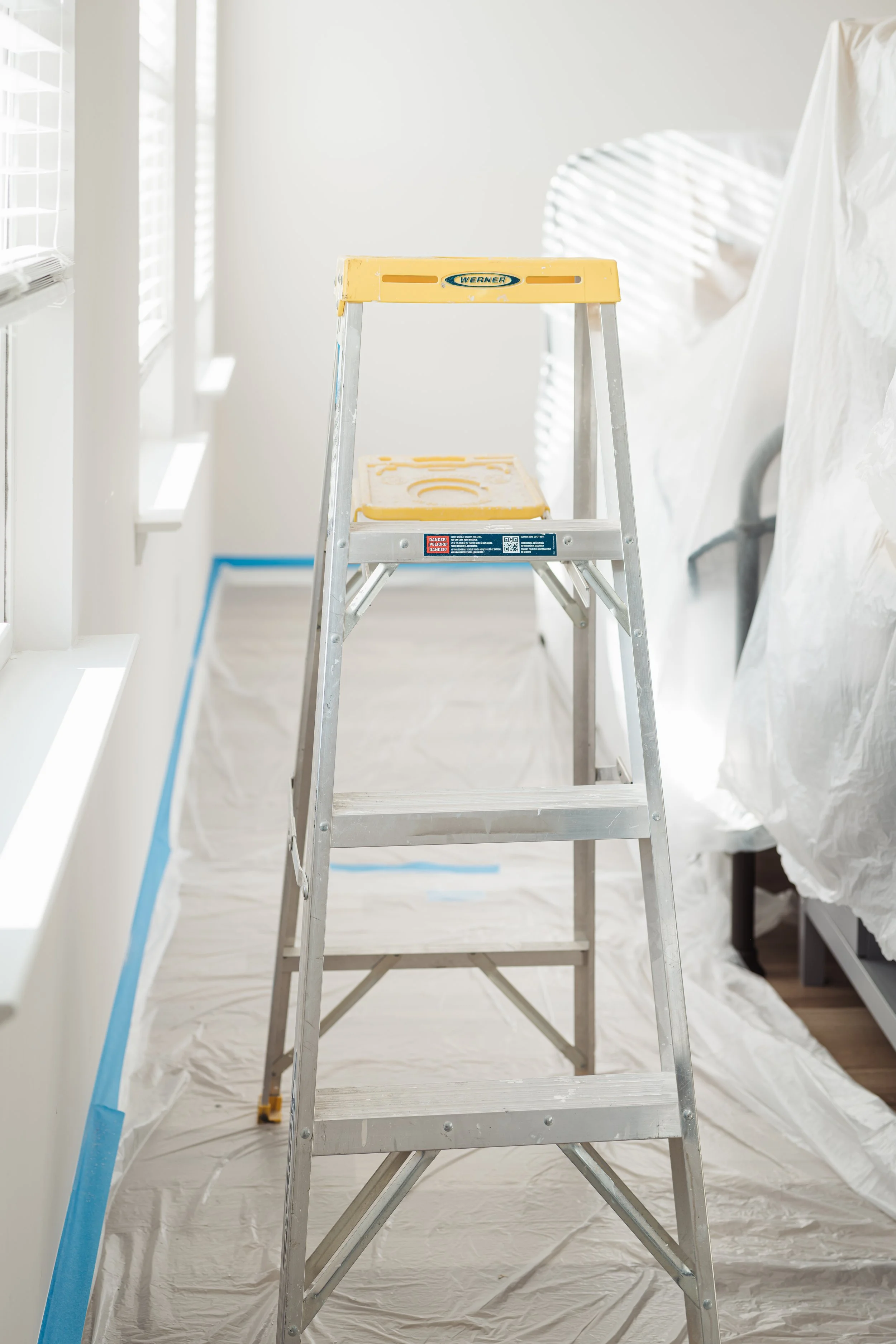 A step ladder in a room with white walls and window blinds, surrounded by painter's plastic sheeting on the floor and walls.