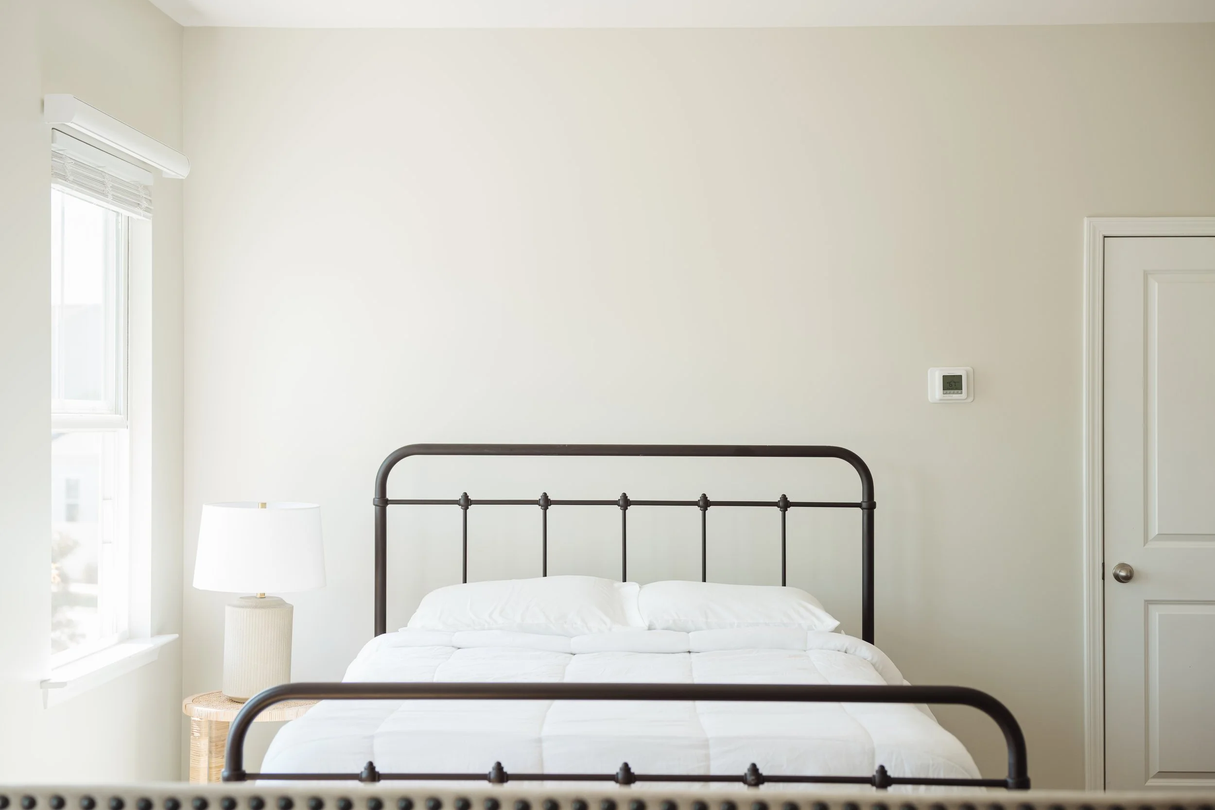 A minimalist bedroom with a metal bed frame, white bedding, a side table with a lamp, and a window with white blinds.