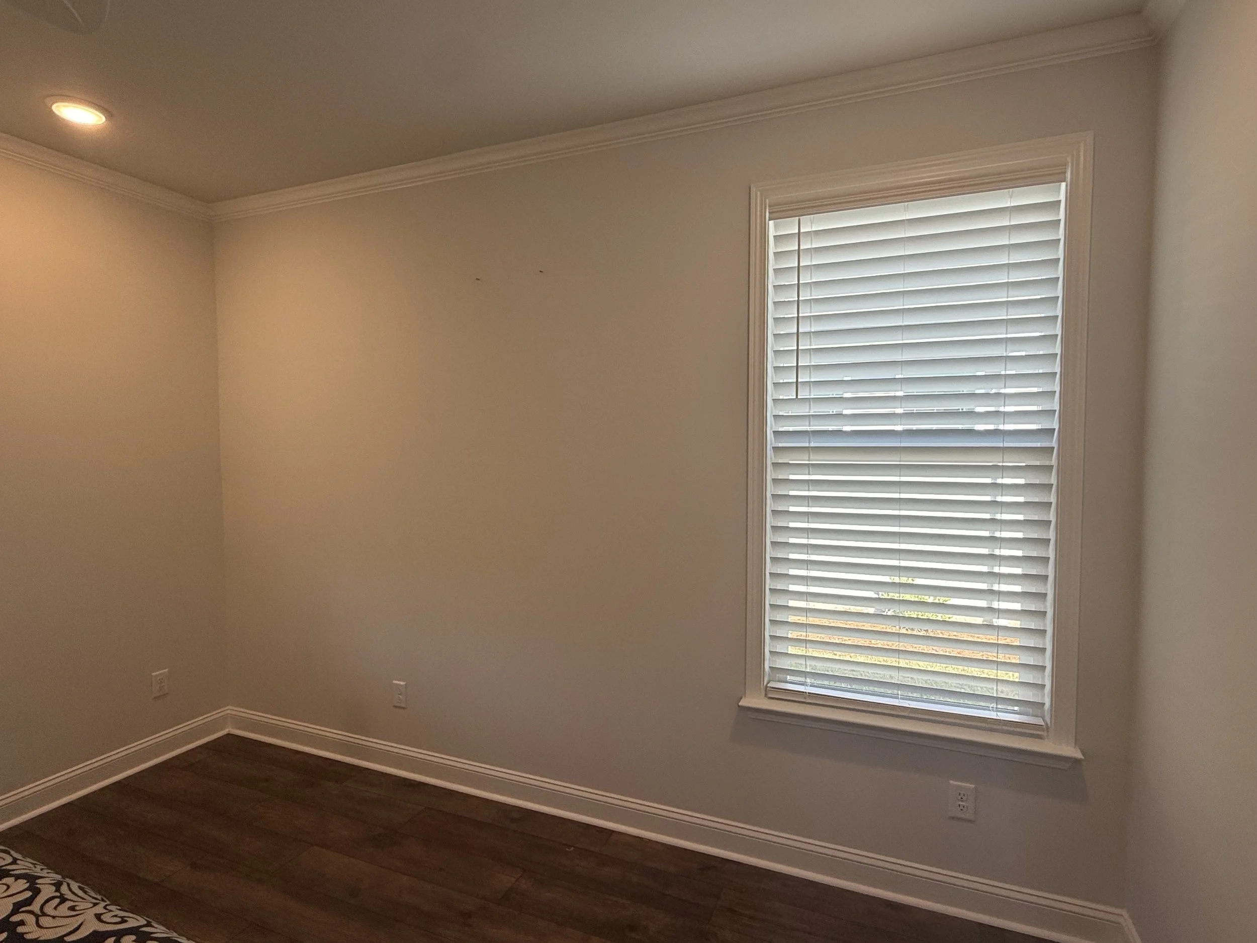 Empty room with beige walls, white ceiling crown molding, hardwood floor, window with white blinds, and recessed ceiling light.