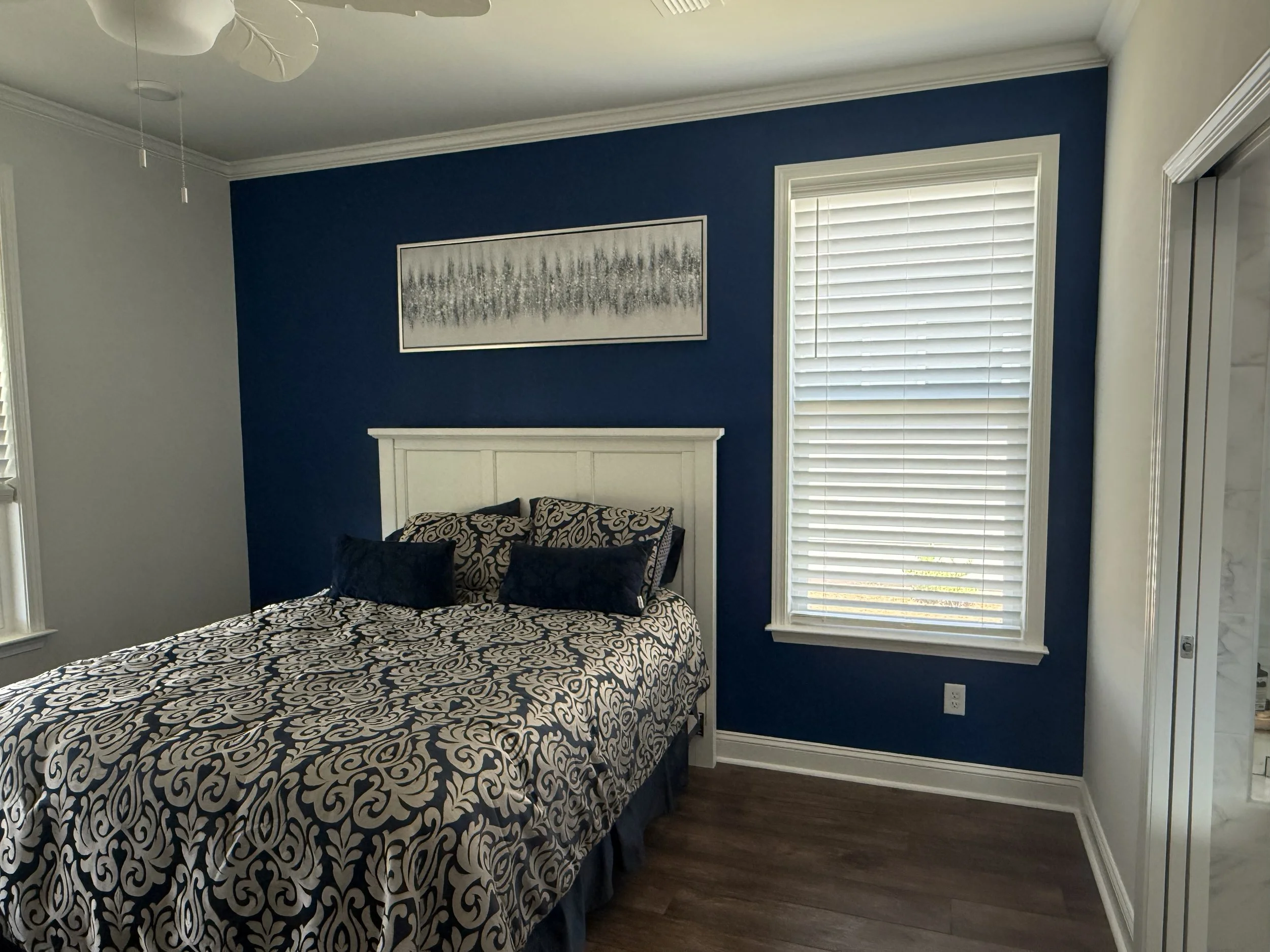 A bedroom with a blue accent wall behind a white headboard, patterned bedding with black and white designs, and a matching pillow. There is a framed black and white artwork above the bed, a window with white blinds to the right, and part of a white c