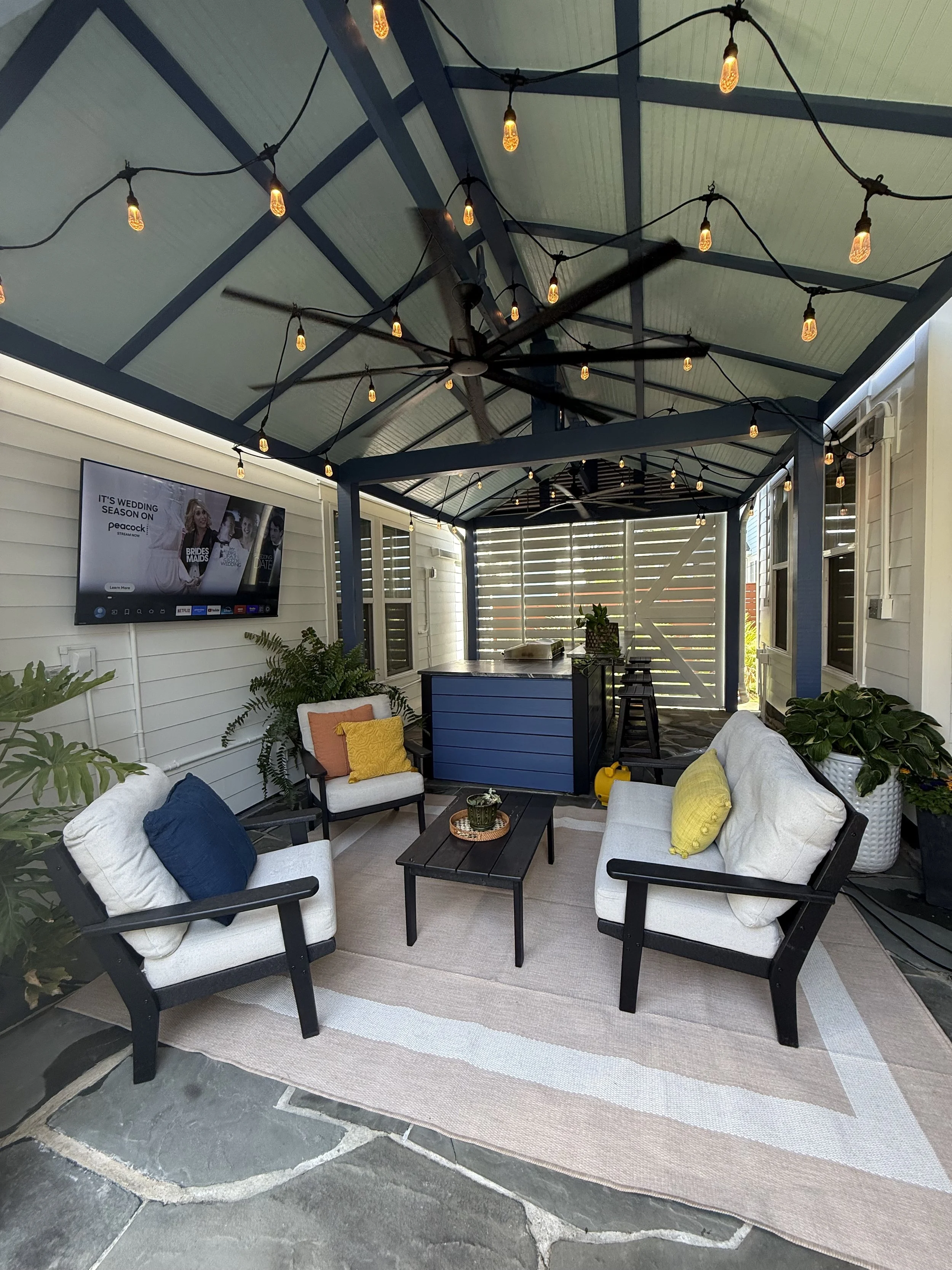 An outdoor patio with string lights hanging from the ceiling beams, white cushioned chairs with blue and yellow pillows, a small black coffee table with a plant, and a wall-mounted TV displaying a streaming service menu. There are plants and a rug on