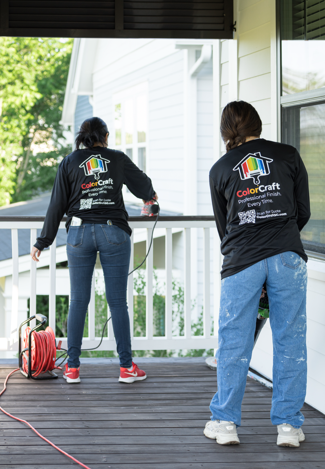 Two women wearing black shirts with the ColorCraft logo power washing a porch on a residential house, with trees and neighboring houses in the background.
