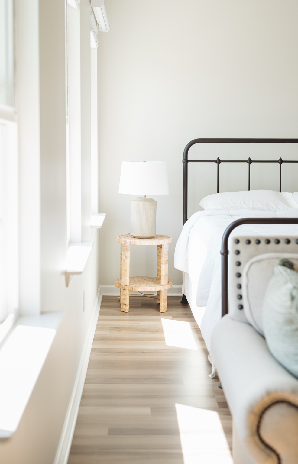 A bedroom with white walls, a bed with white bedding and a black metal frame, a beige bedside table holding a white lamp, and a wooden floor with sunlight coming through the windows.